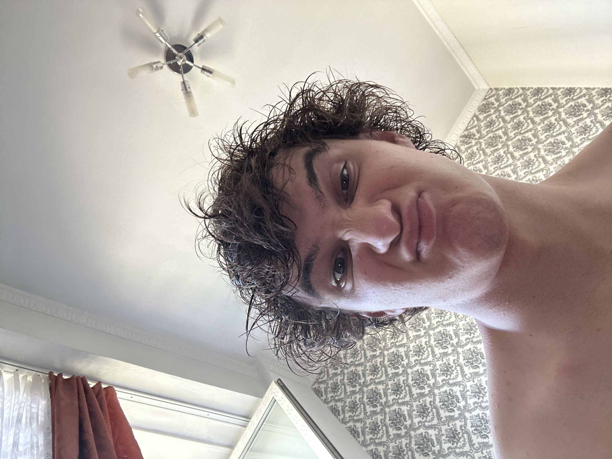 A close-up, low-angle selfie shows a young person with messy, dark, curly hair making a pouting face. The background includes a textured wallpaper, a ceiling with a light fixture, and a window with folded curtains. The image does not contain any identifiable landmarks or contextual information that would allow for determination of its location within Krasnoyarsk, Russia.