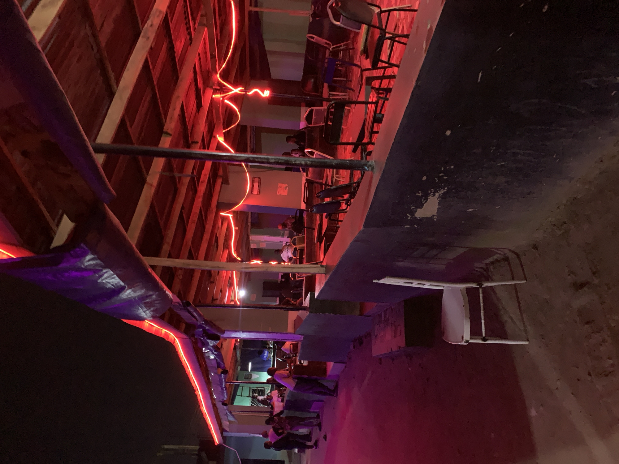 A nighttime scene at an outdoor venue in New Stadium, Nigeria, reveals a group of people gathered near an illuminated structure. The structure, possibly a bar or entertainment area, is constructed with wooden beams and adorned with red neon lights. A dark tarp or covering is partially visible on one side. Several people are standing and interacting near the base of the structure, while others sit at tables inside. Chairs are visible both within the illuminated area and outside, near a concrete wall. The overall environment is dimly lit, with the red neon lights providing the primary source of illumination.