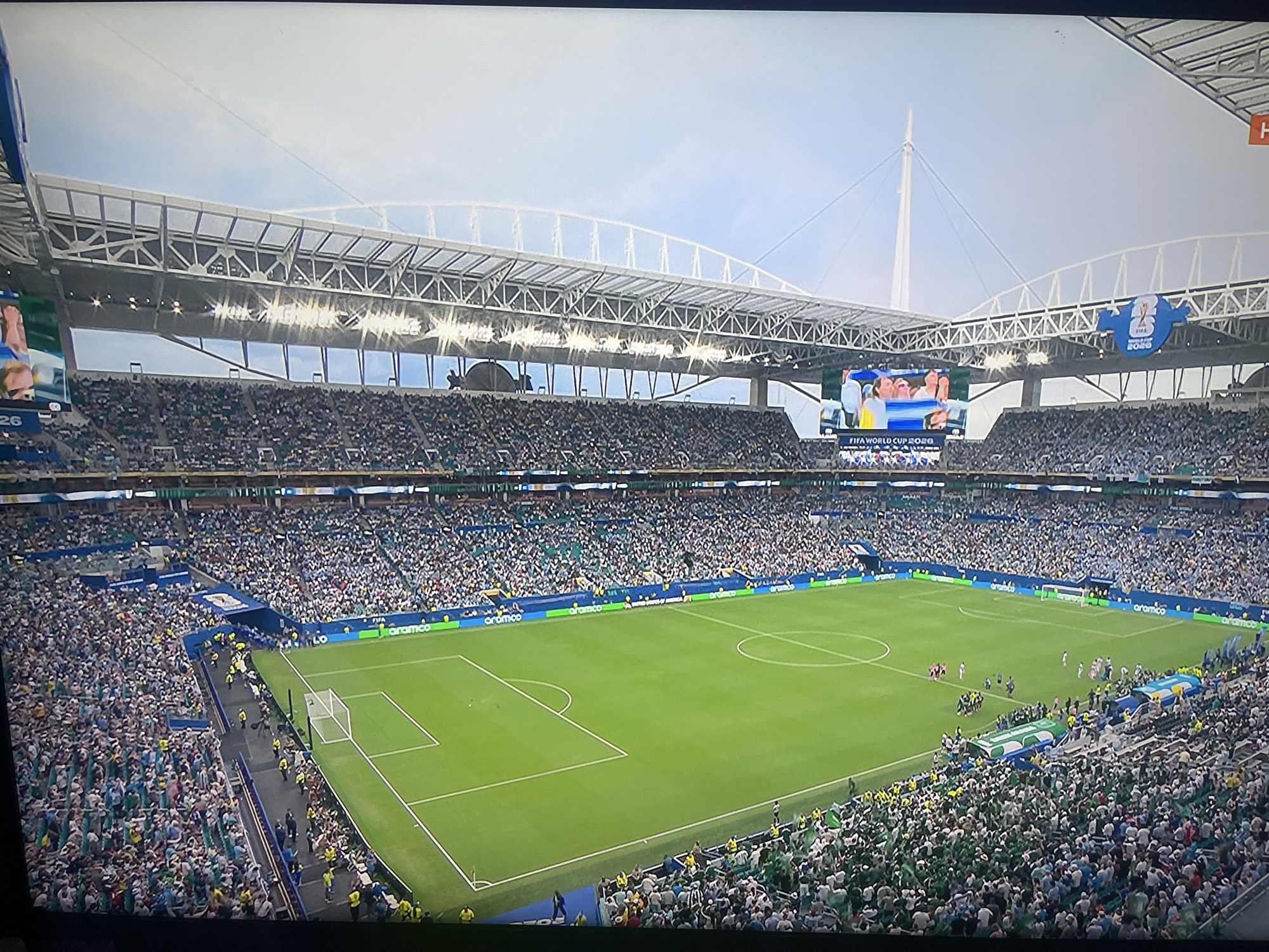 The image depicts a panoramic view of a soccer stadium during a match, filled with spectators. The stadium features a large, modern roof structure and numerous bright lights. A large screen displays images of players, and banners with "FIFA WORLD CUP 2020" are visible. The field is green, with white markings indicating the boundaries and penalty areas. The stands are packed with a diverse crowd wearing predominantly green and white attire. People can be seen in various locations within the stadium, including the stands, the field of play, and along the sidelines. The environment appears to be daytime, with a bright, cloudy sky. The image is a photograph taken from an elevated perspective, likely from another section of the stadium or an aerial view.
It is important to note that the provided image does not depict the city of Ruse, Bulgaria. The context of the "FIFA WORLD CUP 2020" and the stadium's architecture are inconsistent with any known locations in Ruse, Bulgaria.