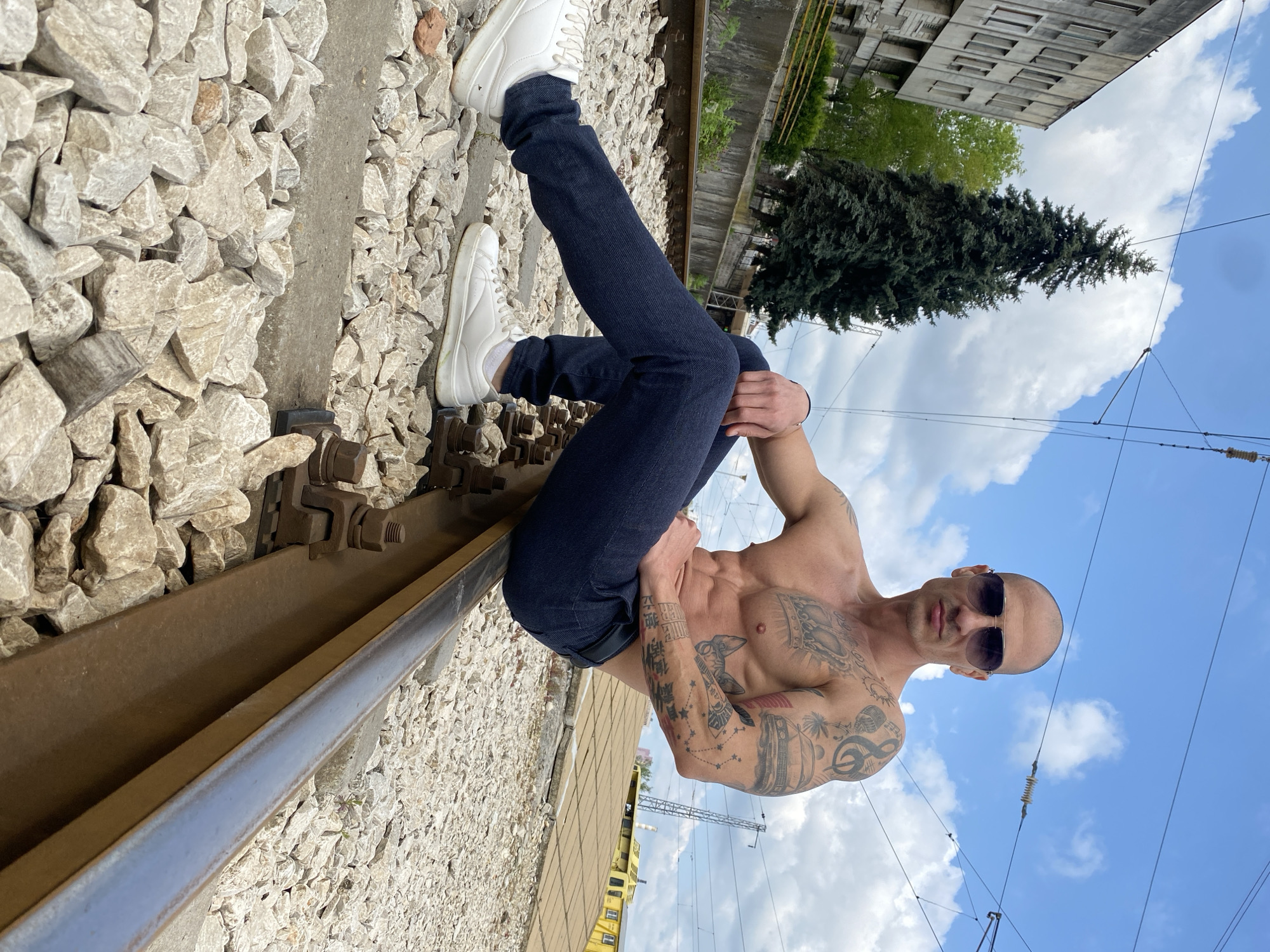 The image shows a shirtless man with tattoos wearing jeans and white shoes, sitting on a railroad track. He is also wearing sunglasses. The location appears to be near a building and a pine tree with a cloudy blue sky in the background. The surrounding area consists of rocks and railroad components. While I cannot positively identify the city in Bulgaria as Pleven, this matches the general environmental characteristics.