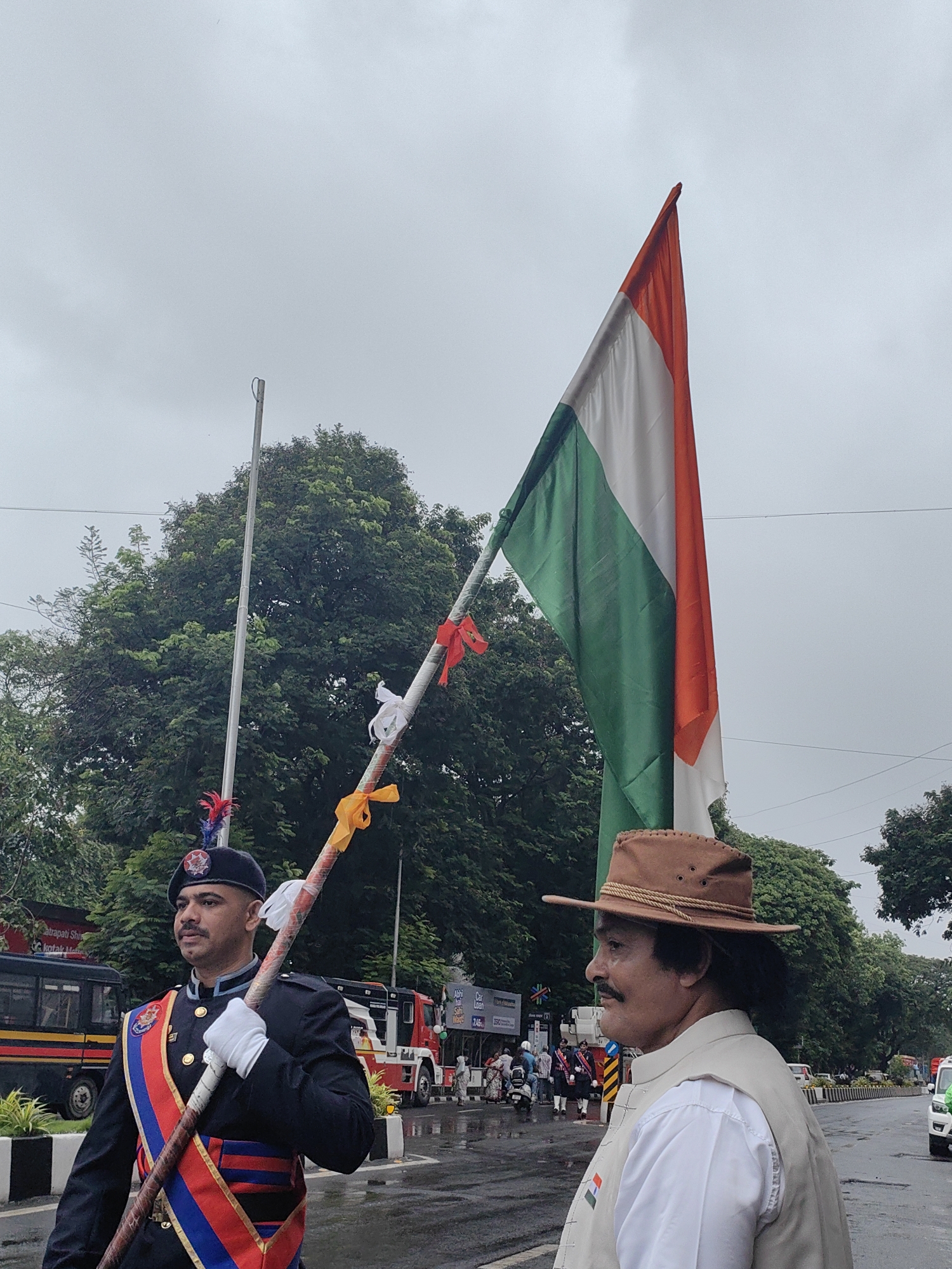 The image captures a street scene in Mumbai, India, likely during a parade or public event. In the foreground, a man in a military-style uniform with a beret adorned with a feather stands holding a decorated pole. He wears white gloves and a sash with red, blue, and gold stripes. To his right, another man wearing a light brown hat and a tan vest over a white shirt is visible in profile, also looking to the side. A large Indian flag, green, white, and orange, is held aloft, partially obscuring the sky.
Behind the individuals, a street is wet, suggesting recent rain, and several vehicles are present, including a red fire truck and a bus. Trees line the street, and the sky is overcast. The overall atmosphere appears to be that of a formal procession or commemoration.