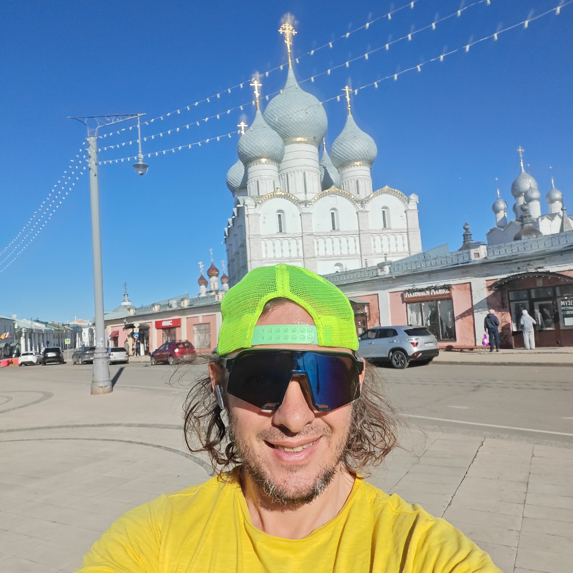 A man is captured in a selfie on a bright, sunny day in Rostov, Russia. He is smiling broadly, wearing a vibrant yellow T-shirt, a neon green mesh cap, and large mirrored sunglasses with blue reflective lenses. His curly brown hair and a beard are visible, and he has wireless earbuds in his ears.
The background features a clear, deep blue sky. Dominating the scene behind him is a large, white architectural complex, likely a Russian Orthodox cathedral, with multiple light grey, onion-shaped domes topped with gleaming golden crosses. String lights are strung horizontally across the sky, adding a festive touch, connecting to a tall street lamp on the left.
The setting appears to be a bustling city street or square. Commercial buildings with varying facades line the street. On a pink-colored building to the right of the cathedral, visible text on a shop sign reads "ПЯТНИЦА ЛАВКА" (Pyatnitsa Lavka - Friday Shop/Stall). Further right, another sign partially shows "МУЗЕЙ" (Museum), suggesting a "Museum of Private Collections." Several cars are parked or driving on the street, and a few pedestrians are visible in the distance. The ground is paved with light-colored flagstones, and shadows are cast by the bright sunlight, indicating daytime. The man's joyful expression suggests he is enjoying the pleasant weather and scenic surroundings.