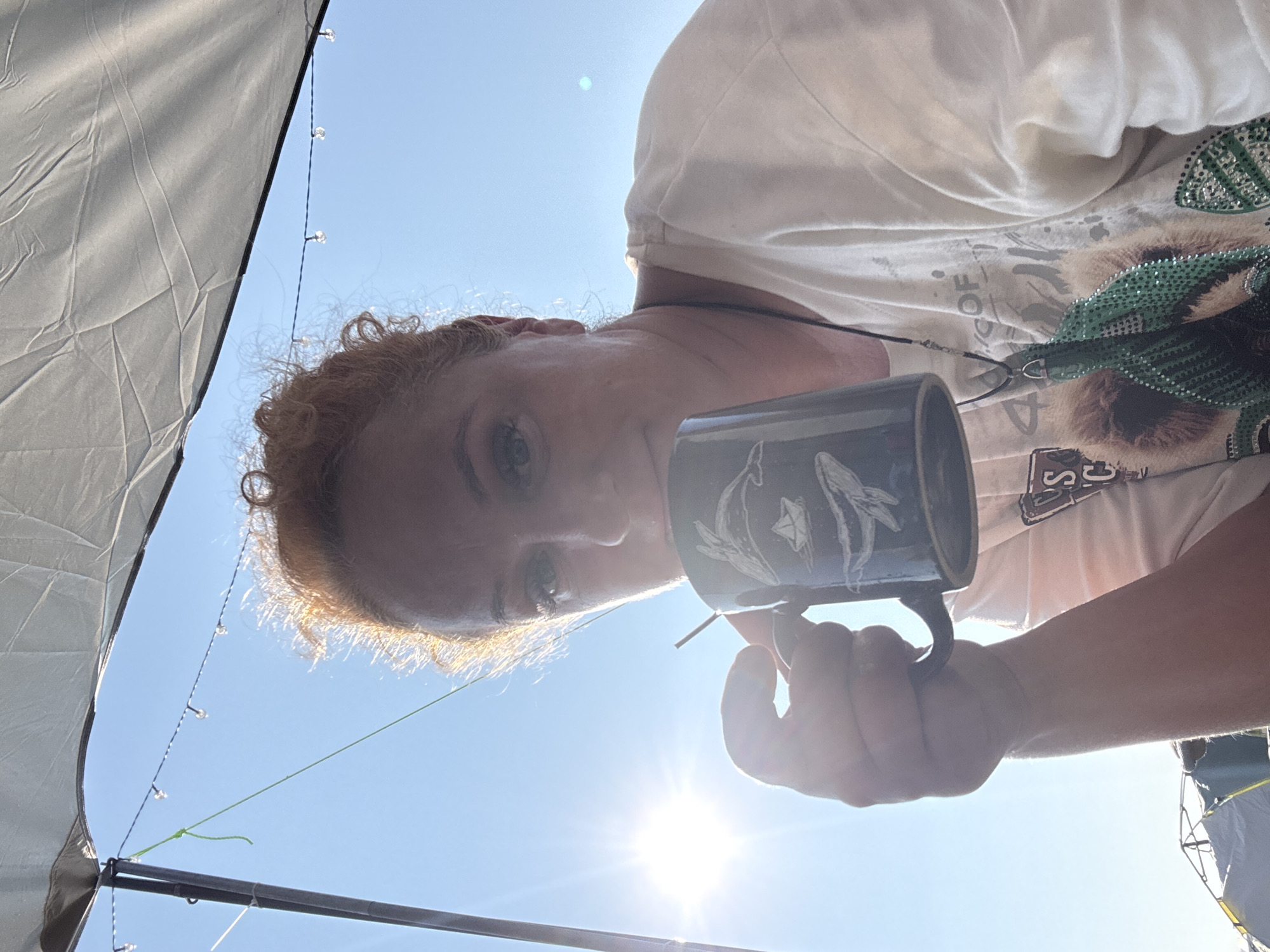 A woman with reddish-orange curly hair is holding a dark blue mug with a design of whales and a sailboat to her lips. She is looking towards the camera with her eyes partially visible. She is wearing a light-colored t-shirt with a graphic print. Behind her, the sky is clear and blue, with the sun shining brightly, causing lens flare. A grey canopy or tent is visible overhead, with some string lights attached to it. Part of a tent structure is visible in the lower left corner. The scene appears to be outdoors, possibly at a campsite.