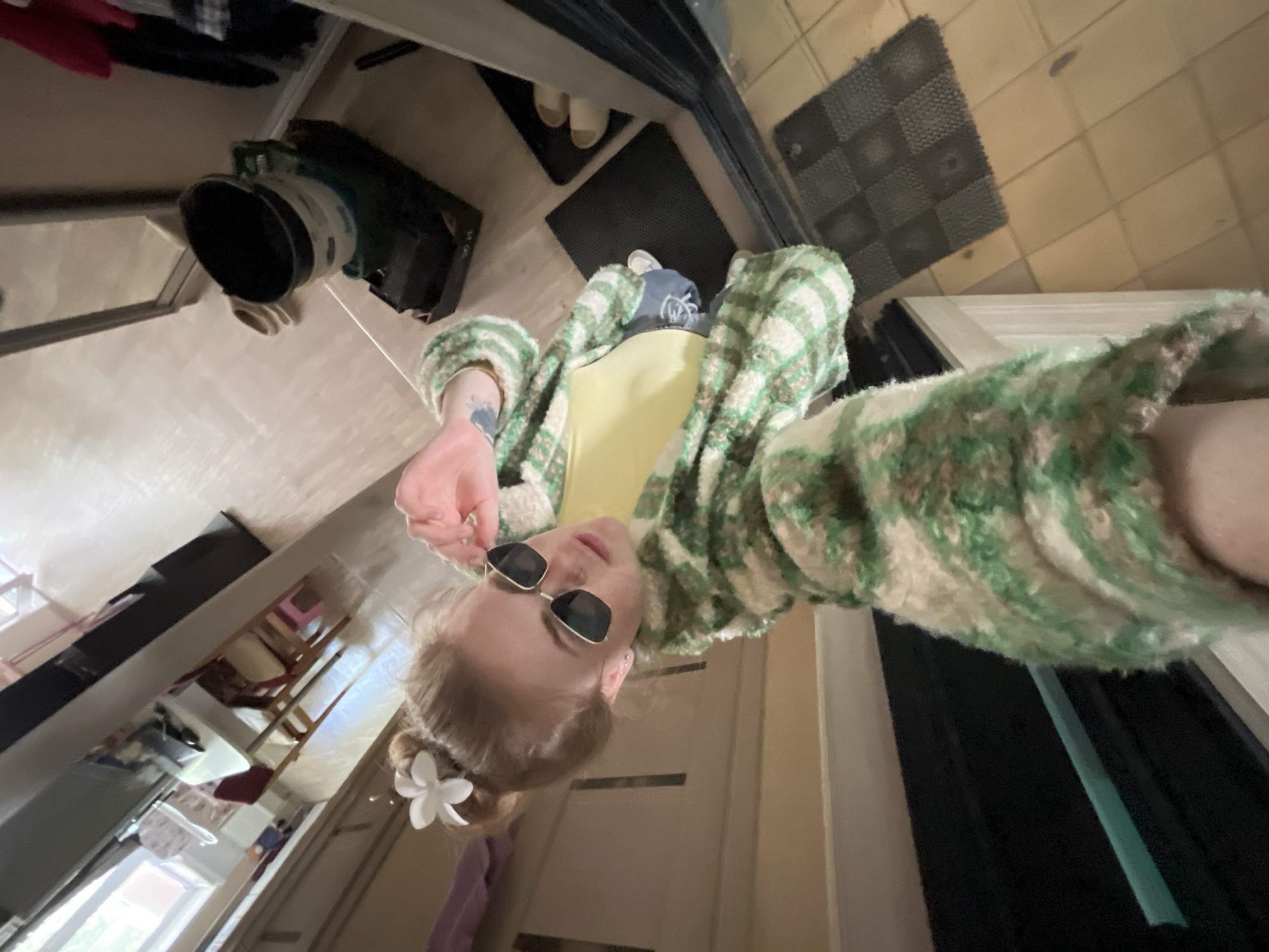 A selfie-style photograph taken from a low angle depicts a fair-skinned individual wearing sunglasses, a light green and cream-colored jacket, and a yellow top. A white flower is visible in their hair.
The surrounding environment includes a kitchen or entryway area. A black object of cylindrical shape is on the left edge of the photograph. A black doormat, and tiles are seen as well.
The location is potentially in Chaikovsky, Russia.
