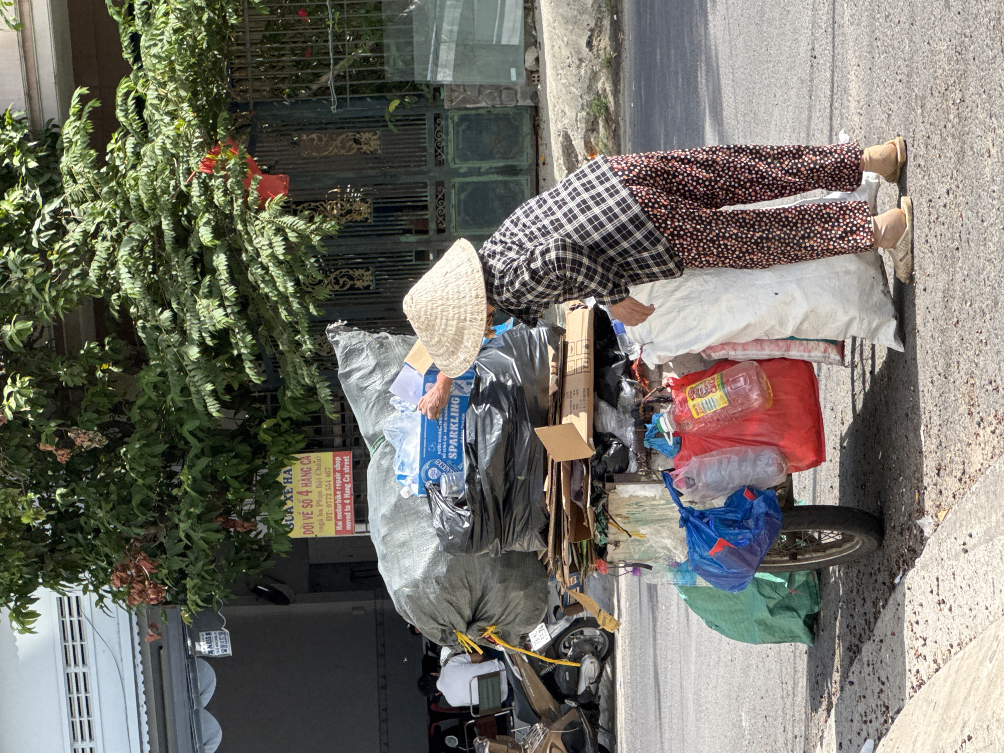 A Vietnamese woman wearing a conical hat and a plaid shirt with patterned pants stands on a street in Nha Trang, Vietnam, next to a cart filled with recyclables. The cart is laden with large bags of plastic and other materials, along with a stack of cardboard boxes and bottles. In the background, a building with a sign in Vietnamese and a metal gate can be seen. A motorcycle is parked on the left side of the image. The scene appears to be a typical street view in a Vietnamese city.