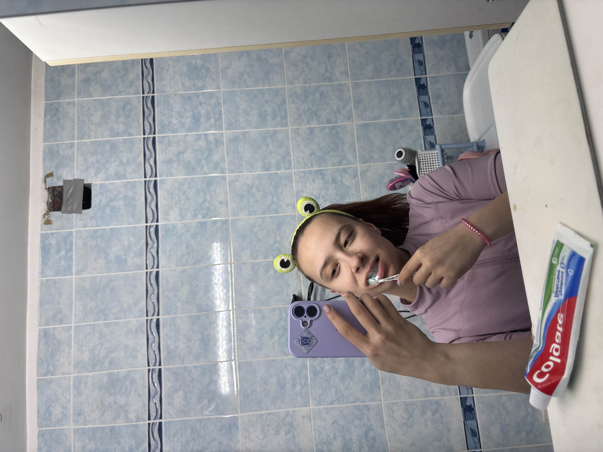 A young woman wearing a headband with frog eyes is brushing her teeth in a bathroom. She is holding a purple smartphone to take a selfie. A tube of Colgate toothpaste sits on a white countertop next to her. The bathroom has blue tile walls with a decorative wavy blue stripe and a toilet visible in the background. There's a rectangular patch covered with duct tape on the wall. Based on available information, the location could be Прикамский, Russia.