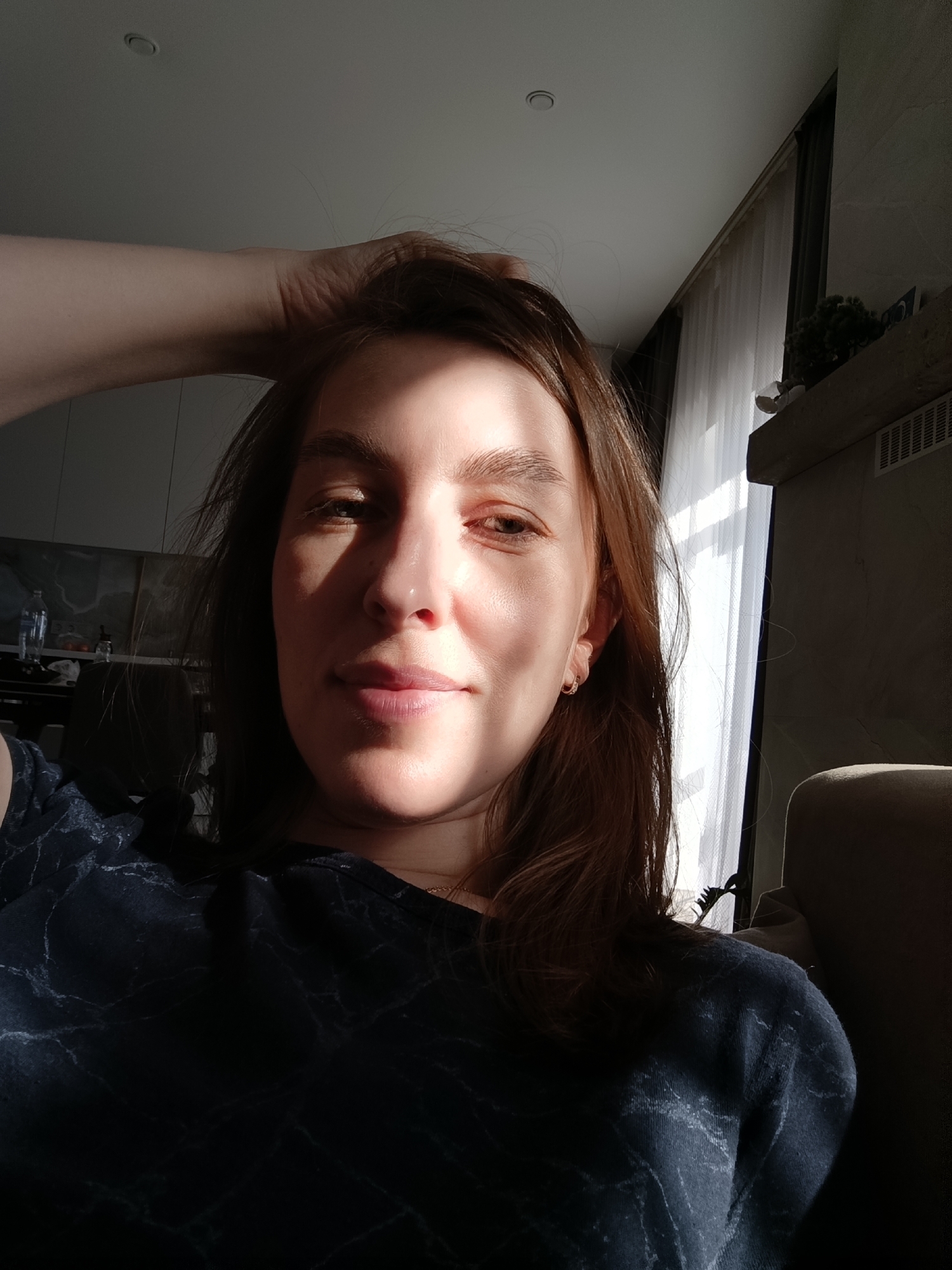 A medium shot captures a fair-skinned woman with shoulder-length auburn hair. She is wearing a dark-colored top with a subtle marble pattern. Her left hand is raised, fingers partially obscuring her hairline. The strong sunlight bathes the left side of her face, casting a shadow on the right. She is wearing small hoop earrings.
Behind her is a partially visible interior. A window draped with sheer white curtains is to her right, with a dark curtain behind it. To the right of the window, there is a shadowed mantelpiece with a plant on it. The background is slightly blurred.
Based on the information provided, it is not possible to definitively confirm that the location is Chaikovsky, Russia.