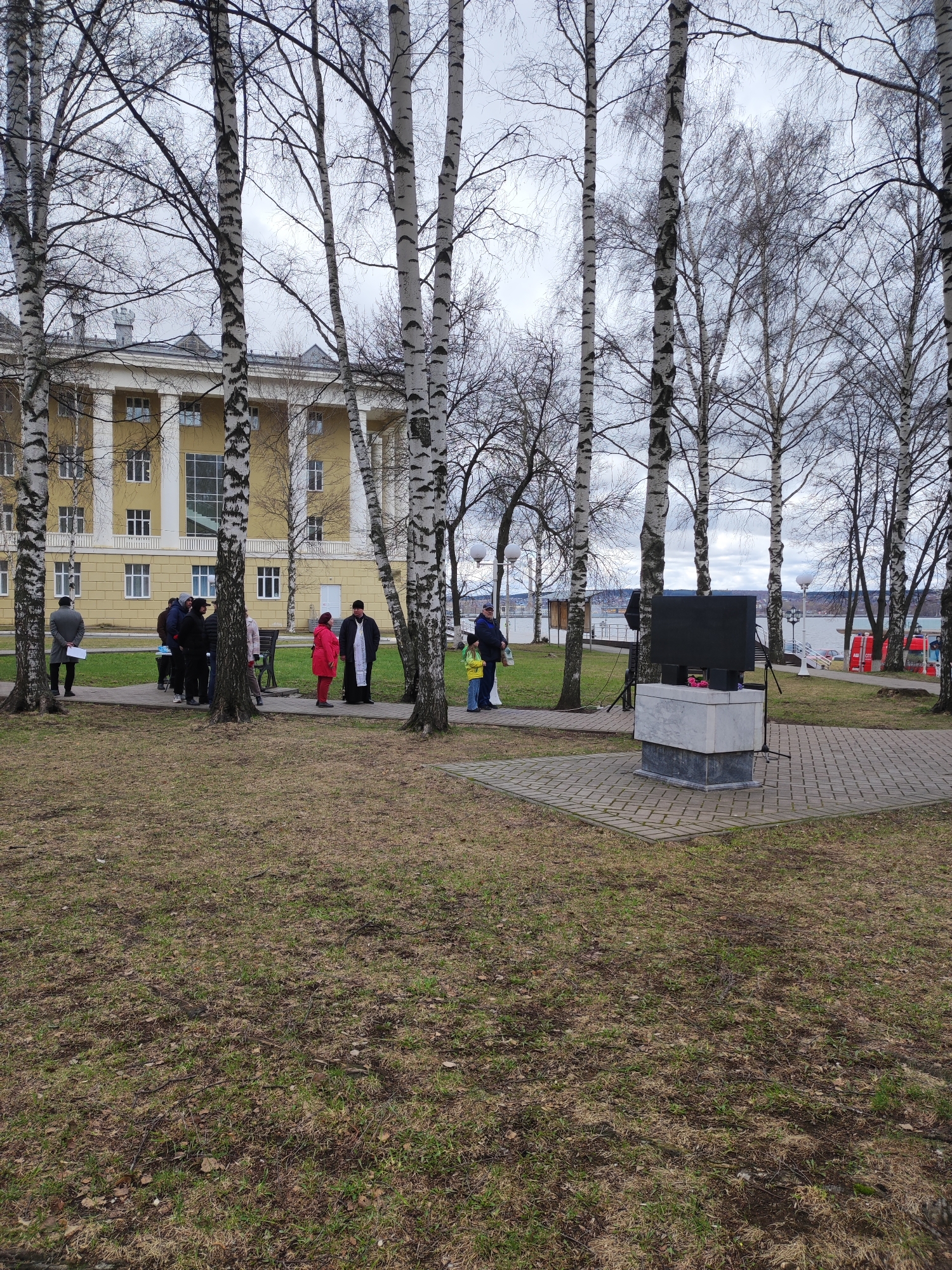 The image captures a gathering in a park setting in Votkinsk, Russia, with a pale yellow building featuring prominent columns in the background. A group of people is standing on a paved walkway. Among them is a person dressed in a black clerical robe with white garments, potentially a priest. A woman in a bright red coat stands out in the group.
A modern-looking monument consisting of stacked marble and dark stone platforms is centrally positioned in the paved area. A large black screen rests on the upper platform.
The foreground features a grassy area with patches of brown leaves. Several birch trees with bare branches stand prominently throughout the scene. In the background, a body of water is visible, partially obscured by the trees. The sky is overcast, suggesting a cloudy day. Red and white emergency vehicles can be seen in the far background.