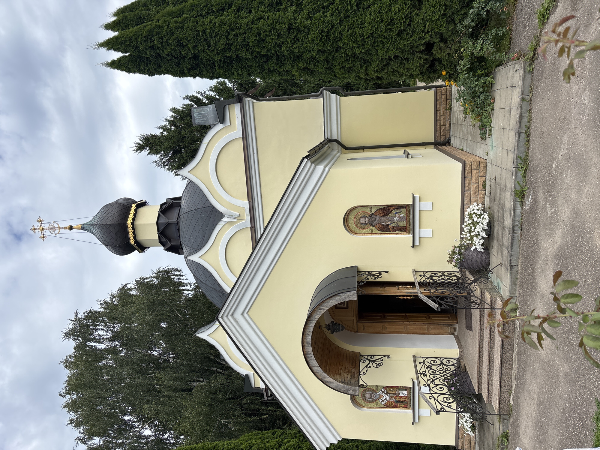 The image shows a small, pale yellow church with a black dome topped with a golden cross. The church is surrounded by trees and has a light-colored stone base. Two mosaic images of religious figures are visible on the facade, flanking the entrance. A set of stone steps leads up to the open wooden doors of the church. Black wrought-iron railings with decorative scrollwork line the steps. Potted plants with white flowers are placed on either side of the entrance. The ground in front of the church is paved with square tiles and some asphalt. The sky is overcast. The church is located in Zvenigorod, Russia.