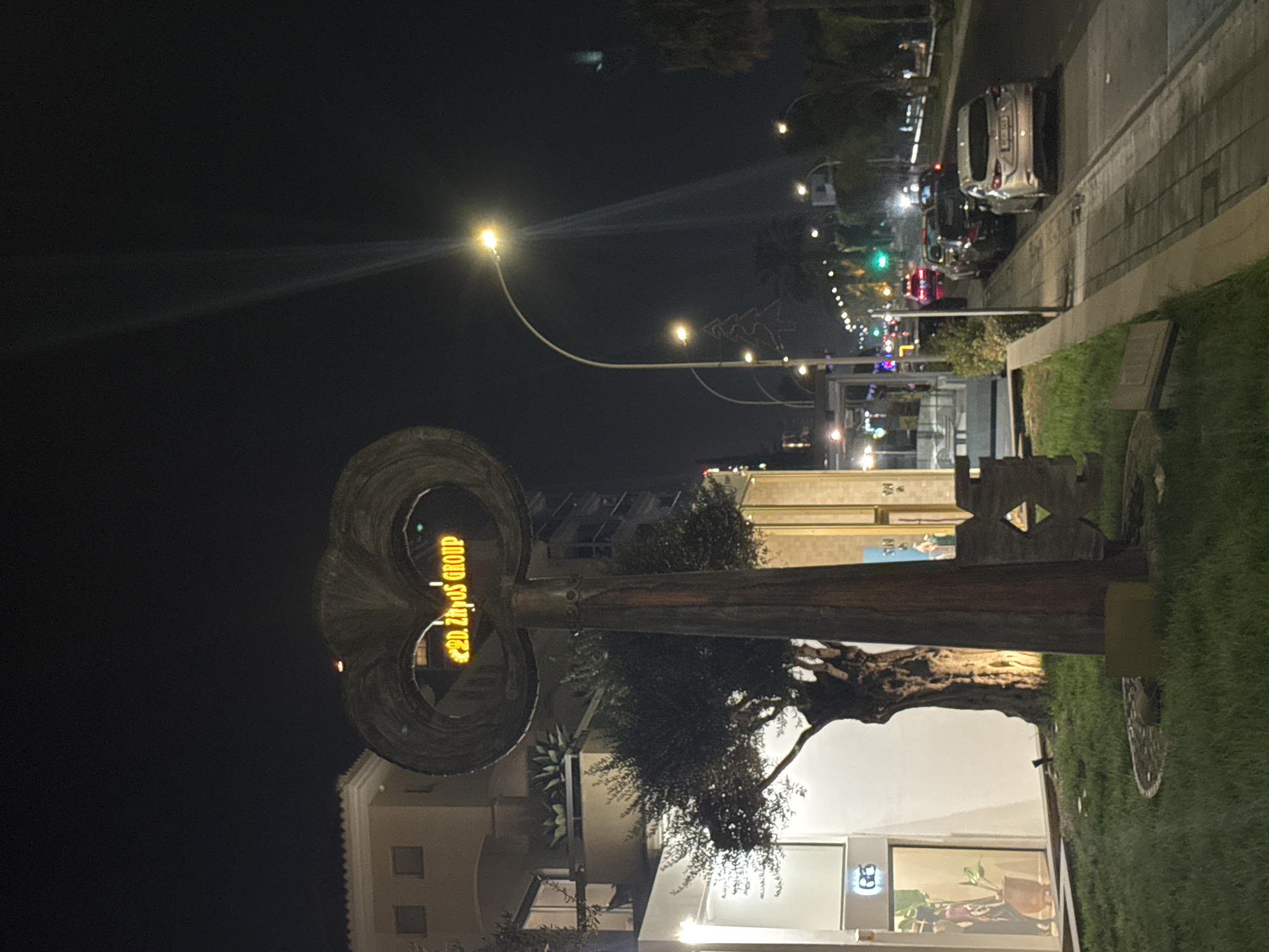 An outdoor nighttime scene depicts a street in Germasogeia Municipality, Cyprus. In the foreground, a large, abstract metal sculpture stands on a patch of grass. The sculpture is illuminated by a yellow LED sign displaying "D. ZAHOS GROUP". To the left of the sculpture is a building with large windows. To the right, a sidewalk runs parallel to a street lined with parked cars and streetlights. A tall, curved streetlight with a bright bulb illuminates the scene. The background shows more buildings, palm trees, and the dark night sky, with some light rays visible, possibly from a distant light source. A Christmas tree outline decoration is visible on a pole in the distance.