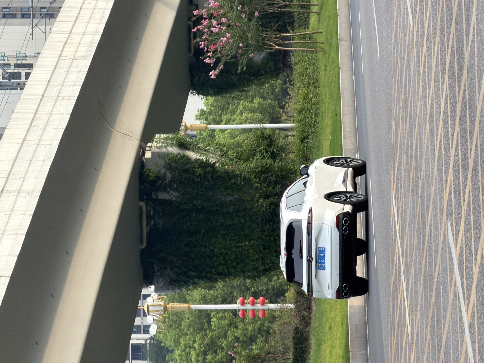 A white SUV is seen from the rear-right at eye-level, driving on a paved road in Jiangjin, China. The vehicle has dual exhaust pipes and a license plate that reads "D 21418". Behind the car, a concrete overpass casts a shadow, and the supports are covered in lush green ivy. To the left of the car, a lamppost with red lanterns is visible, and further in the background, several modern buildings can be seen. The road has yellow grid markings for a pedestrian crossing. The overall environment appears to be a suburban or urban area with ample greenery.