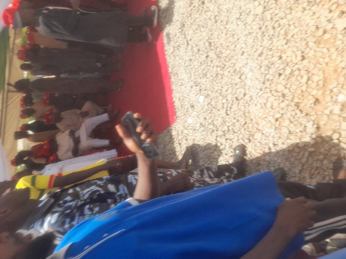 The image shows a group of people standing in a gravel lot in Gusau, Nigeria. The people are standing under a white canopy tent and are arranged in a line. The person in the front is holding a black smartphone. There is a red carpet in front of the people, and it appears they are attending a ceremony or event.