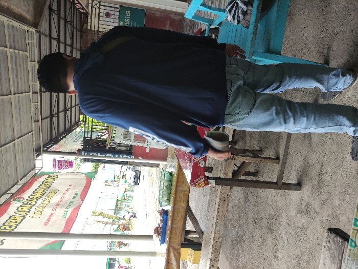 The image captures a daytime scene in Kepanjen, Indonesia, featuring a man standing with his back mostly to the viewer, slightly turned to the left. He is wearing a dark blue long-sleeved shirt, light blue jeans, and dark athletic shoes. His hands are positioned near a simple metal table, and he appears to be holding a dark grey cup or small pot.

The setting is an open-air or semi-outdoor covered area, possibly a casual eatery or warung, with a concrete floor and a roof supported by metal structures. To the right, a bright blue wooden bench or chair is visible. In the background, there are other blurred figures seated at tables.

Notable details include:
- A large banner on the left background displays partially visible text: "SEGER REK..." (Indonesian slang for "fresh, man/dude...") and "YAMARA SS", "15 WARUN APL". Crucially, "Jalan Baru Kepanjen" is clearly readable, confirming the location.
- A green sign in the upper right background shows "UM FUSTISI!", possibly related to a university or organization.
- On the table in the foreground, a red object, possibly a wrapper or container, has the word "BAKSO" printed on it, indicating a popular Indonesian meatball dish.
The overall ambiance is relaxed and casual, typical of a local dining spot.