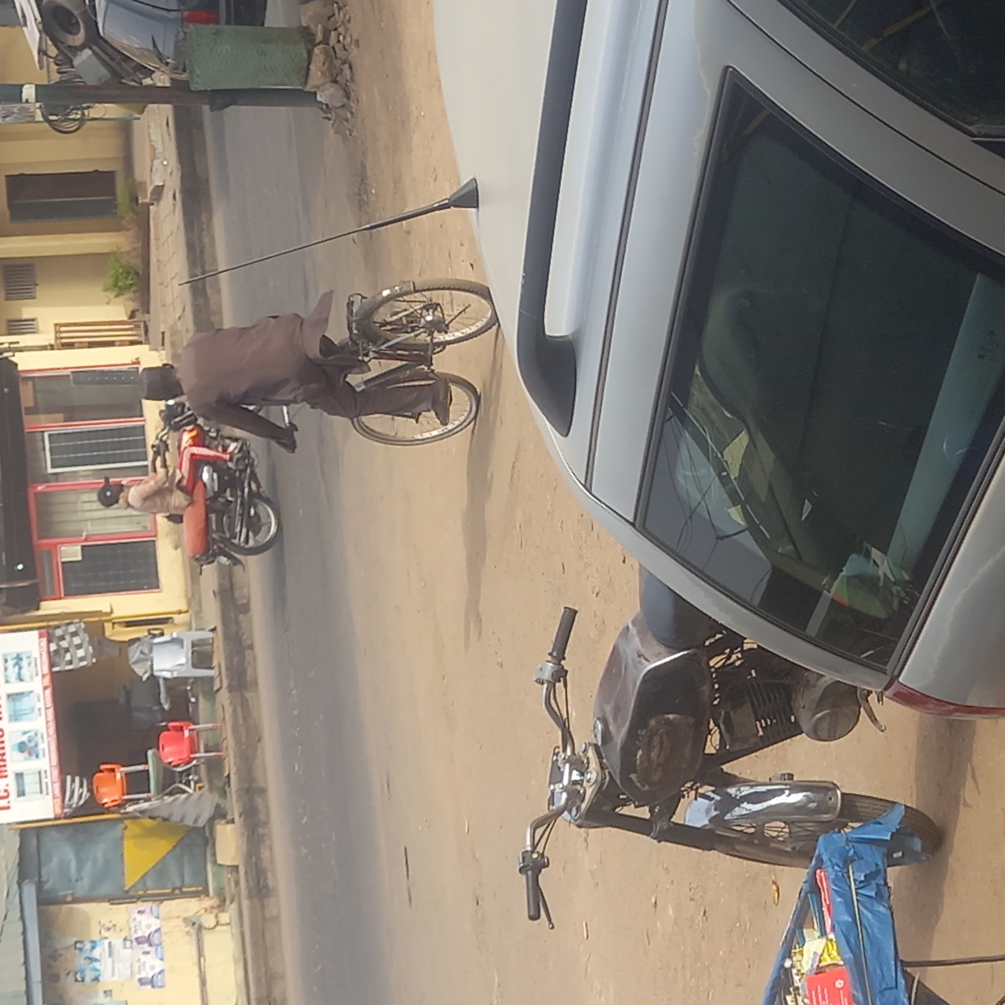 This image depicts a vibrant street scene in Dadi Riba, Nigeria, captured during daylight hours on a clear, sunny day. The perspective is slightly elevated, looking across a street that features both a paved road and a dirt shoulder.
In the immediate foreground on the right, a significant portion of a silver or white car is visible, showcasing its side profile, windows, and roofline. Below the car, on the dusty ground, a black motorcycle with prominent chrome handlebars and exhaust is parked, facing left. A blue tarp or bundled item rests near its front wheel.
Central to the mid-ground, a man dressed in a flowing brown garment is actively engaged with a dark-colored bicycle, appearing to be working on or inspecting it. The bicycle is situated on the dirt part of the street.
Further into the background on the left, another man, wearing light-colored clothing and a cap, is seated on a red and black motorcycle. This vehicle is parked near a light yellow building with red-framed windows and doors. A commercial sign on this building is partially visible, displaying "T.C. MANDU INT" along with images suggesting household items or furniture. Two brightly colored chairs, one orange and one red, are placed outside the shop, adding a splash of color to the scene. The street itself is paved, indicating an active thoroughfare. The overall impression is one of typical daily life and commerce along a busy local street.