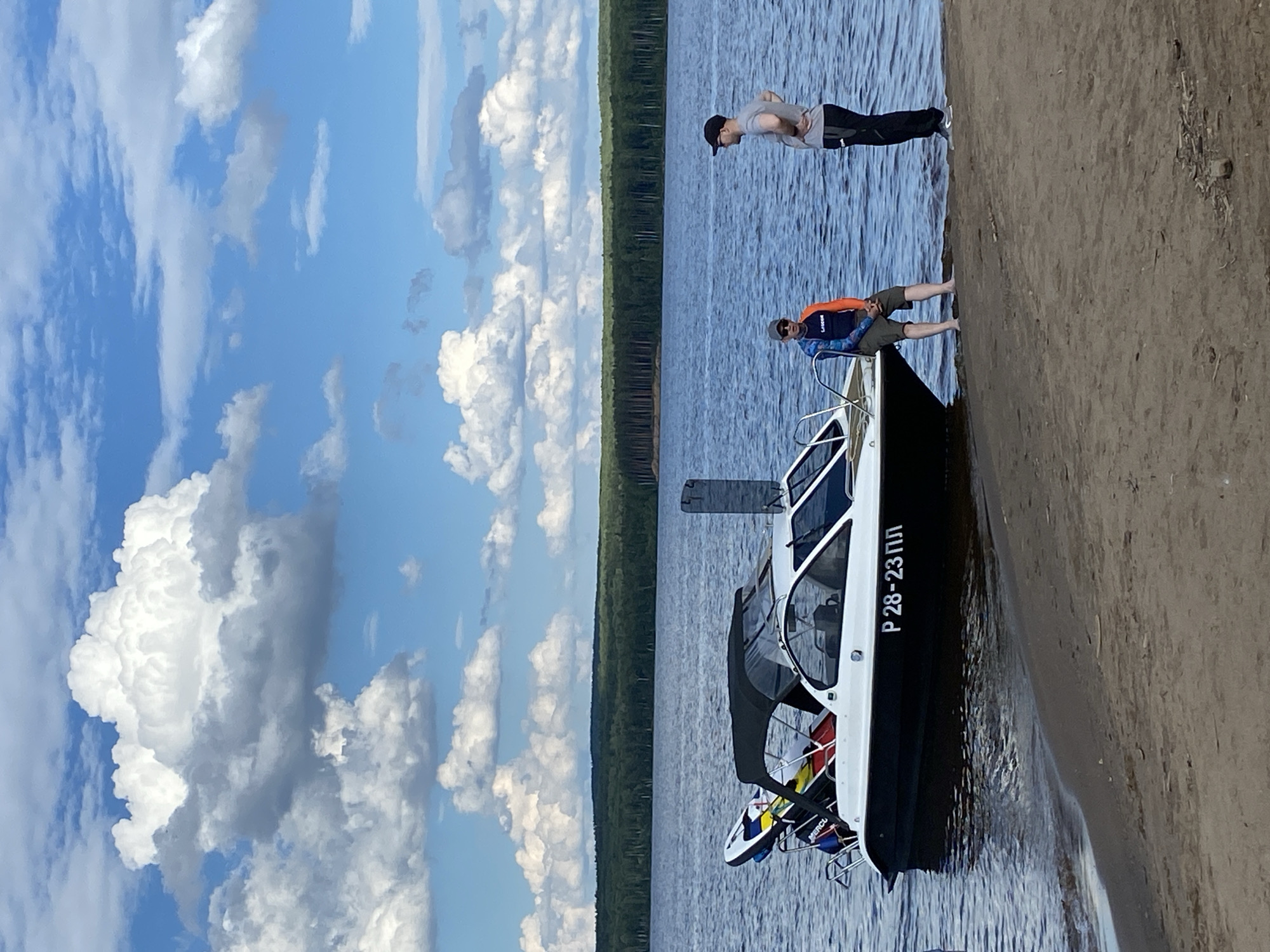 In Chaikovsky, Russia, on a sunny day with a partly cloudy sky, a motorboat is moored near a sandy shore. Two men are present. One man, wearing a colorful long-sleeved shirt and shorts, is seated on the edge of the boat's hull. The other man, in a gray t-shirt and black pants, stands on the shore next to him. The boat, identified by the registration number "Р 28-23 ПЛ" on its side, has a black hull and a white cabin. Several water sports equipment items, including what appear to be surfboards or paddleboards, are secured to the boat's stern. The background features a calm blue body of water leading to a dense forest that lines the distant horizon.