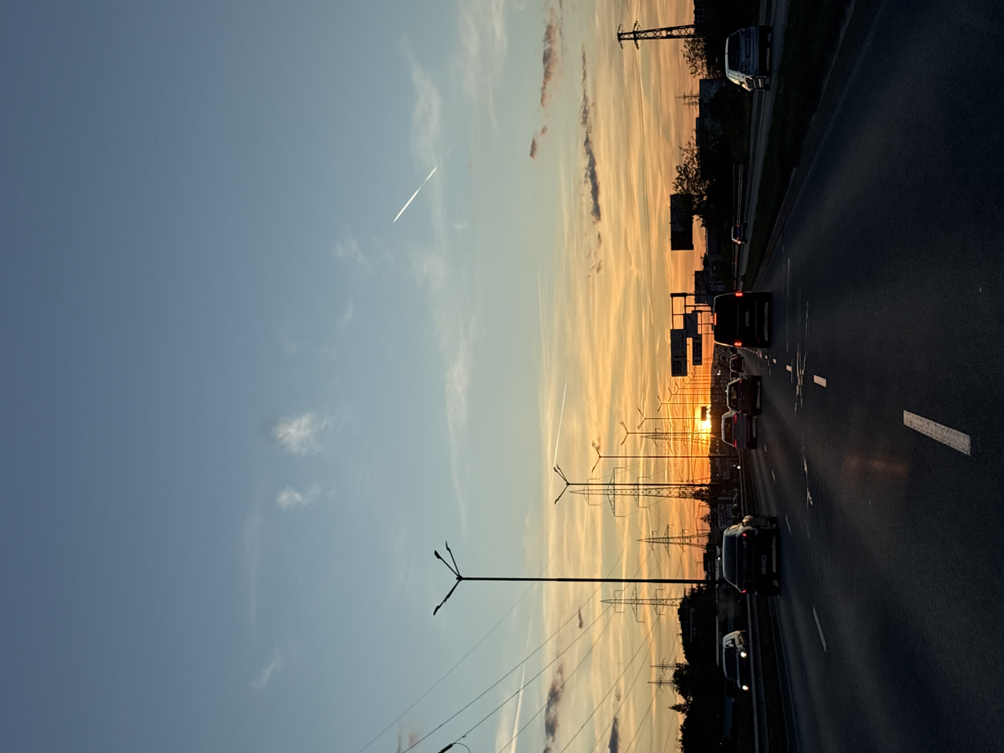 The image depicts a highway at sunset, likely in Sofia, Bulgaria, due to the architectural style of the lampposts and power line pylons visible. Cars are traveling on the right side of the road. The sky transitions from blue to a warm orange hue as the sun sets. Cloud cover is present. Power lines and streetlights are visible along the side of the road. There is a contrail in the sky.