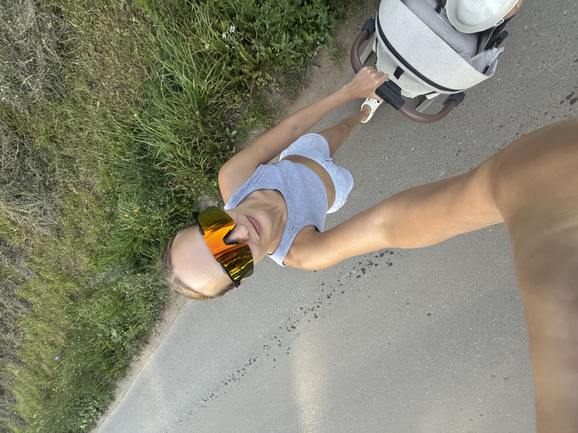 A woman is walking with a stroller on a paved road. She is wearing a grey crop top, grey shorts, white Crocs, and oversized sunglasses with orange lenses. The stroller is light gray with a brown handle. To the right of the road is a grassy embankment. The scene is filmed from a high angle, giving the impression of looking down on the woman and stroller. There is no specific indication of the location being in Podshivalovo, Russia.
