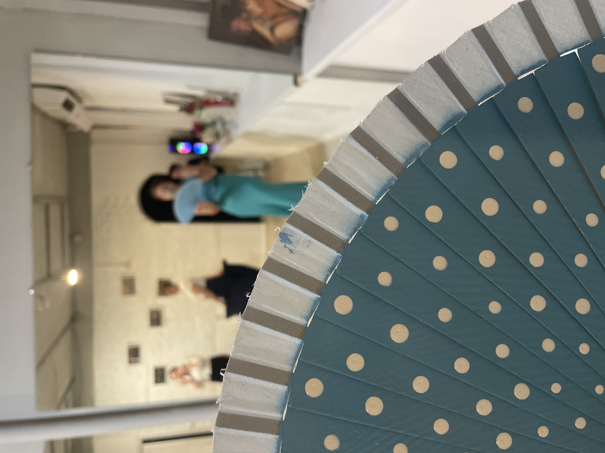 The image shows a close-up of a teal fan with cream polka dots, angled towards the viewer. The fan is partially open, revealing pleats. The background is blurred but appears to be an indoor event or gathering. Two women are visible in the mid-ground, one on the left wearing a white top and black bottoms, and another on the right wearing a white top and dark bottoms. Further back, a third woman in a blue dress is holding a light blue fan. The environment suggests an art exhibition or a social event, with framed pictures on the wall and a counter with items on it. The location is specified as Sochi, Russia, but there are no visual cues in the image to confirm this.