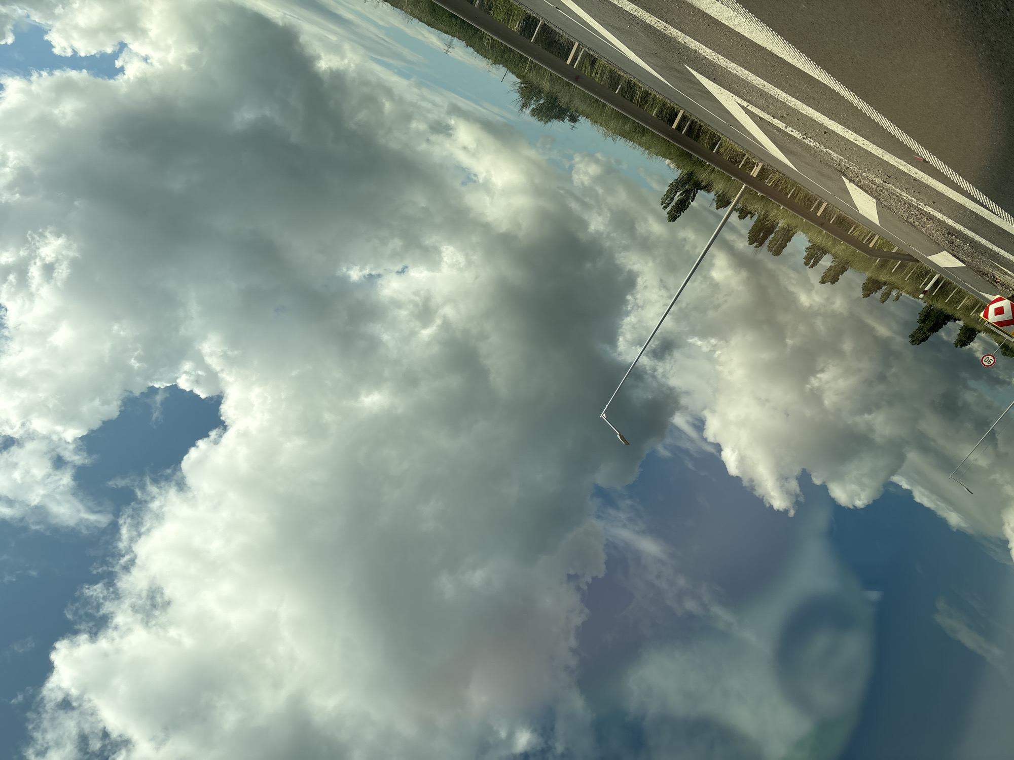 The image, rotated 90 degrees clockwise, captures a view primarily of the sky and clouds, seemingly from inside a vehicle looking upwards and outwards.
The dominant feature is a vast expanse of clouds, varying in density and color. Large, fluffy white and grey cumulus clouds fill much of the frame, with patches of clear blue sky visible between them. The sunlight indicates daytime, though the presence of darker grey clouds suggests either a partially overcast day or approaching weather.
In what would be the upper right portion of the correctly oriented image, a section of a highway or major road is visible. It features distinct white road markings, including solid lines, dashed lines, and arrows, indicating lanes and directions. The road appears to be asphalt. Along the roadside, there is a strip of green vegetation, including grass and trees, reflecting in the glass. A thin grey streetlight or lamp post extends from the lower left towards the upper right, partially obscured by the clouds.
Two road signs are visible on the right side of the road. One is a red-bordered, white diamond-shaped sign, which typically indicates a priority road. Below it, a circular speed limit sign displays "90" in black on a white background with a red border. Directly beneath the speed limit sign, the numbers "06" are visible, likely part of a road marker or additional information.
In the bottom right corner of the image, a faint, blurred reflection of a person's face or head is visible on the glass surface through which the photo was taken, suggesting the photographer was inside a car or bus. The overall scene is consistent with a drive through a rural or semi-rural area near Hrabarsko, Bulgaria, given the European style of the road signs.