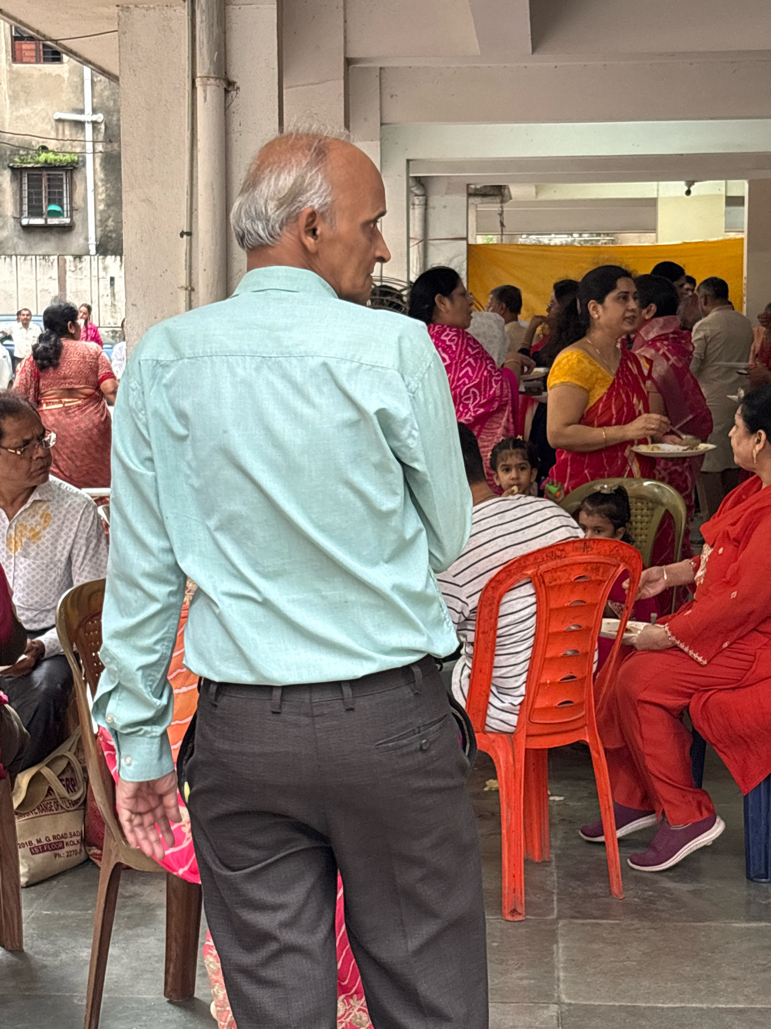 The image depicts an outdoor gathering of people, likely a social or religious event, in Howrah, India. In the foreground, a bald man with grey hair is seen from behind, wearing a light blue collared shirt and dark grey trousers. He is facing away from the camera, towards a group of people.
To the left, partially visible, is a man wearing glasses and a white patterned shirt, seated in a wooden chair. A beige canvas bag with text in English and a phone number is on the floor next to him.
In the mid-ground and background, several individuals are engaged in conversation and eating. A woman in a red and pink sari is holding a plate and a utensil, appearing to be eating. A group of children are also present. Two young girls are visible, one looking towards the camera. Other adults, dressed in traditional Indian attire like saris and kurtas, are mingling. An orange plastic chair is prominently placed in the mid-ground. The environment suggests an open-air venue, possibly adjacent to a building with concrete pillars and textured walls. A yellow tarp is visible in the background, suggesting a shaded area. The ground appears to be paved with large stone tiles.