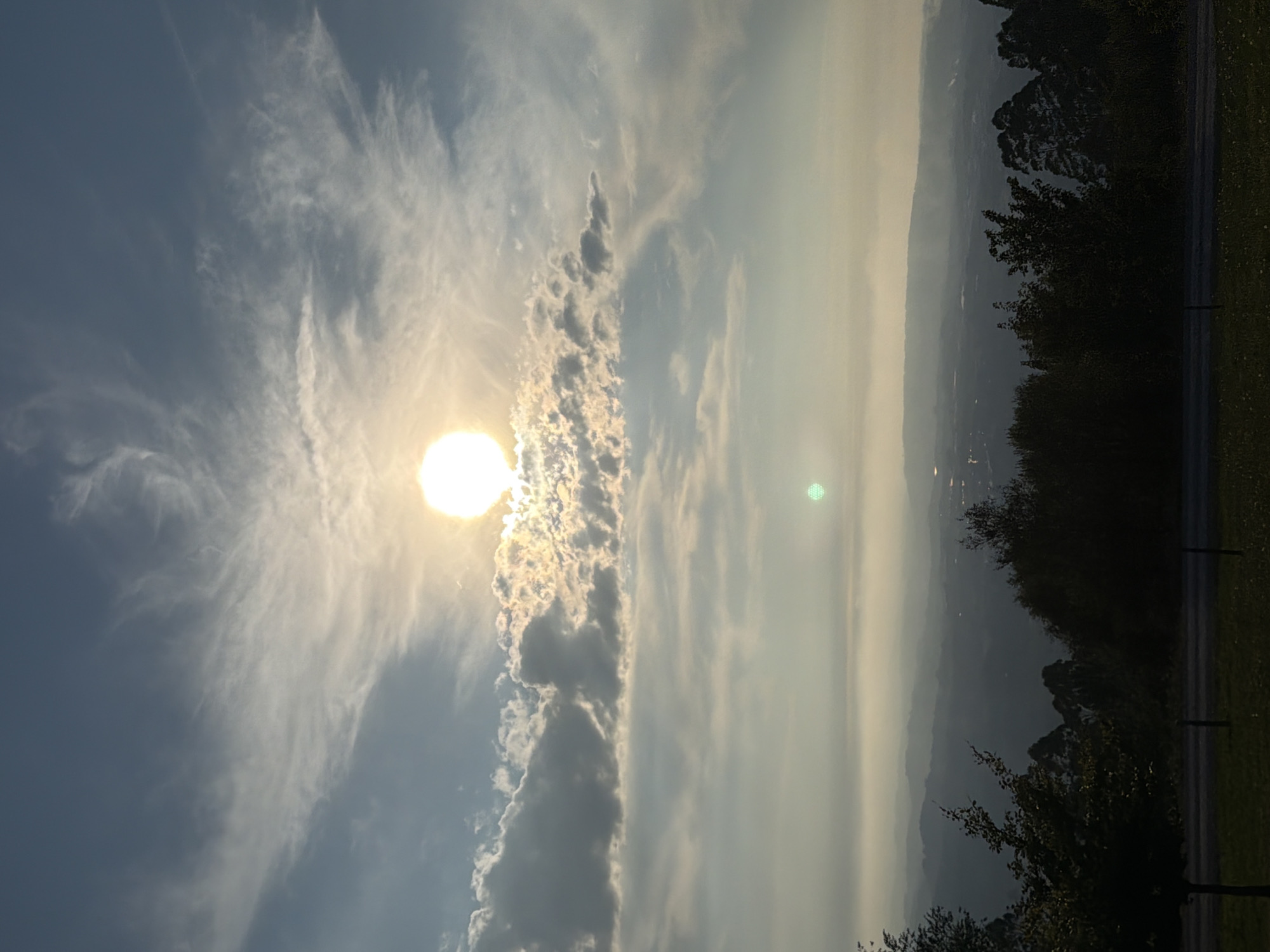 The image captures a panoramic view from a high vantage point, likely overlooking a valley or coastal region. The sky dominates the upper half of the frame, with the sun shining brightly through a scattering of wispy clouds. The sun appears as a luminous orb, casting a warm glow that highlights the textures of the clouds. Below the sky, layers of hills recede into the distance, shrouded in a soft, atmospheric haze that gives the landscape a sense of depth and mystery. In the foreground, the silhouettes of trees are visible, with dark foliage contrasting against the lighter tones of the sky and landscape. The trees are situated on a grassy slope, suggesting a natural, elevated setting. The overall impression is one of tranquility and natural beauty, possibly during a late afternoon or early morning. The specific location in Gijón, Spain, is not identifiable from the visual information provided in the image.