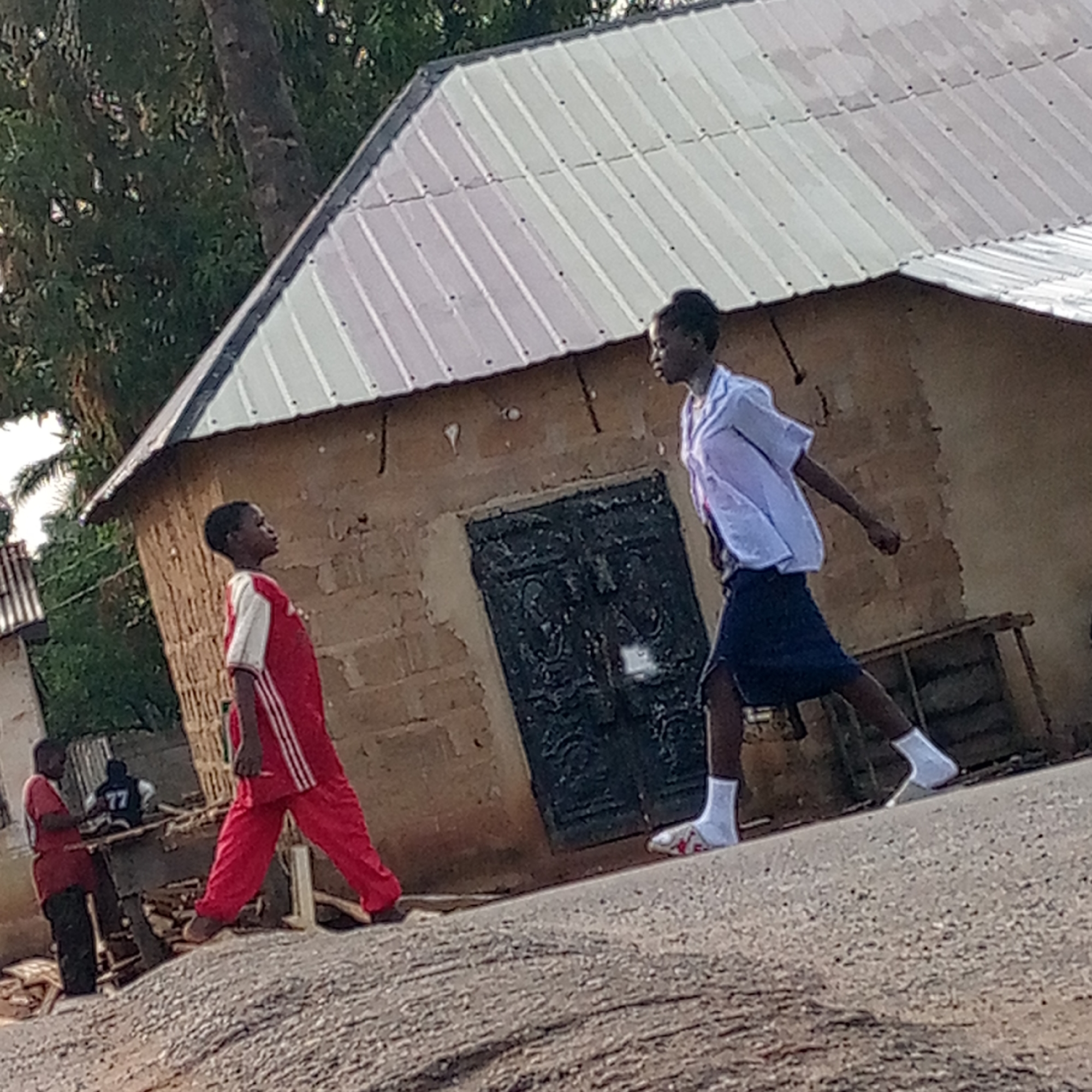 The image shows two children in Mararaba, Nigeria. One child, wearing a red tracksuit with white stripes, is walking uphill. The other, dressed in a white short-sleeved shirt, dark skirt, and white socks and sneakers, is walking downhill. They appear to be looking at each other. Behind them is a small building with a metal roof and a dark, ornate door. In the background, two men are standing near a makeshift table, one of them wearing a jersey with the number 77. The ground appears to be unpaved. Vegetation and parts of other buildings are visible in the background.