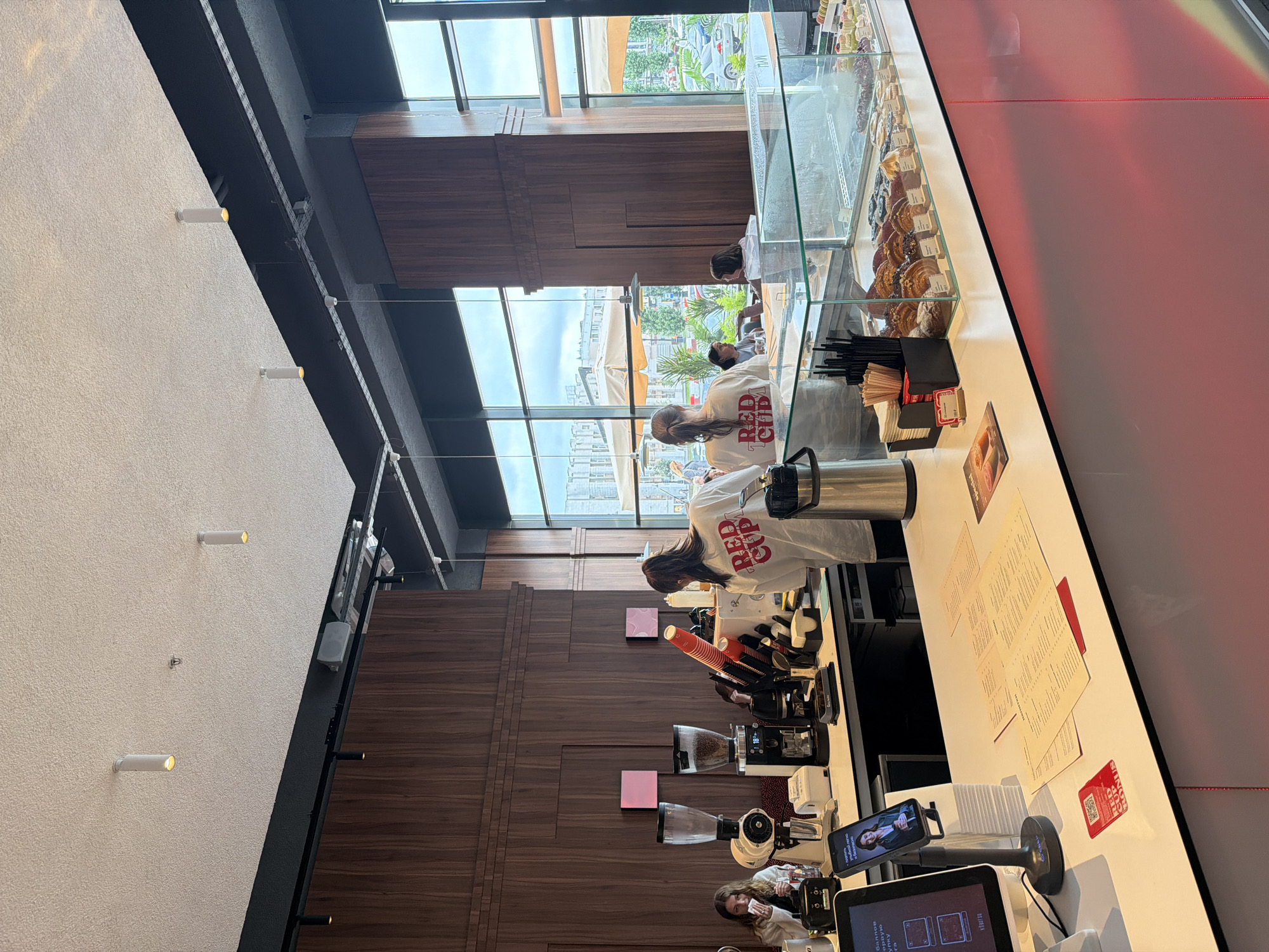 The image depicts the interior of a modern coffee shop, likely located in the city of Perm, Russia, given the presence of Cyrillic text on menus and the style of the establishment. The scene is a busy counter area with several individuals. Two baristas, wearing white t-shirts with "RED CUP" printed in red on the back, are actively working. One barista is facing away from the camera, serving or preparing something behind the counter. The other barista is also behind the counter, facing towards the viewer and looking to the left.
In the background, a female customer is seated at a table, looking at her phone. Another person, possibly a customer, is also seated at a table in the background, partially obscured. The counter itself is well-stocked with a variety of pastries displayed in a refrigerated glass case. There are also coffee machines, grinders, cups, and other service equipment. The decor features dark wood paneling on the walls and a bright, clean counter surface. Large windows offer a view of an urban outdoor setting with trees, buildings, and parked cars, suggesting it is daytime. Overhead lighting fixtures are visible on the ceiling. A digital menu or ordering screen is mounted on a stand at the front of the counter.