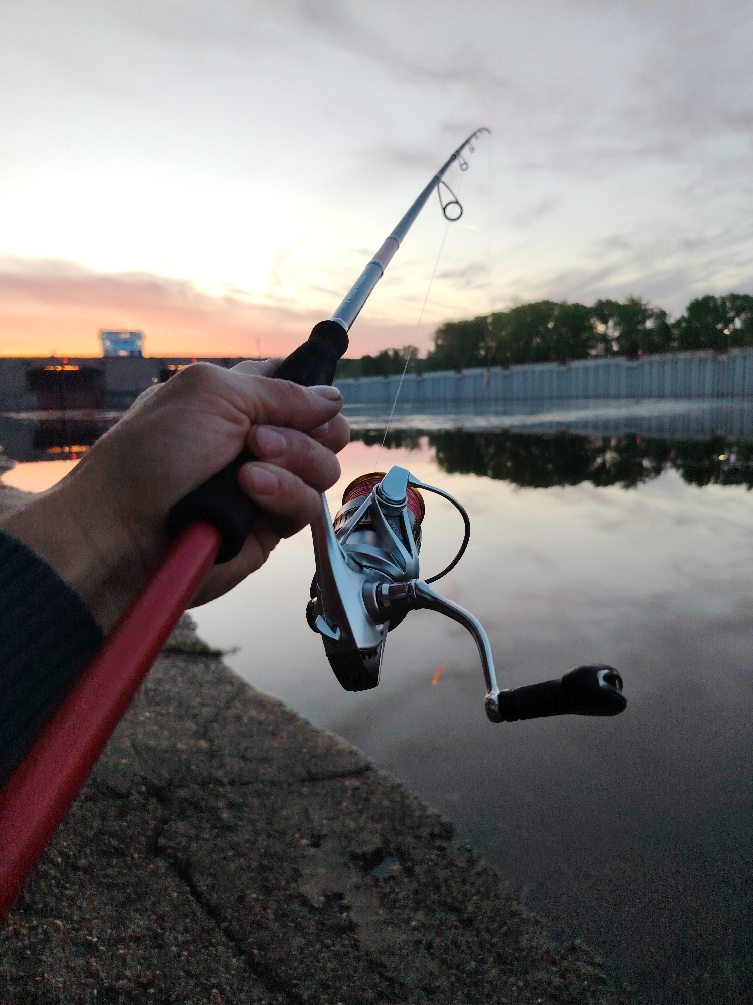 A first-person view shows a hand holding a red and gray fishing rod with a silver spinning reel at dusk. The background features water reflecting the pink and orange sky. In the distance, there's a concrete structure and a line of trees. The location may be Chaikovsky, Russia.