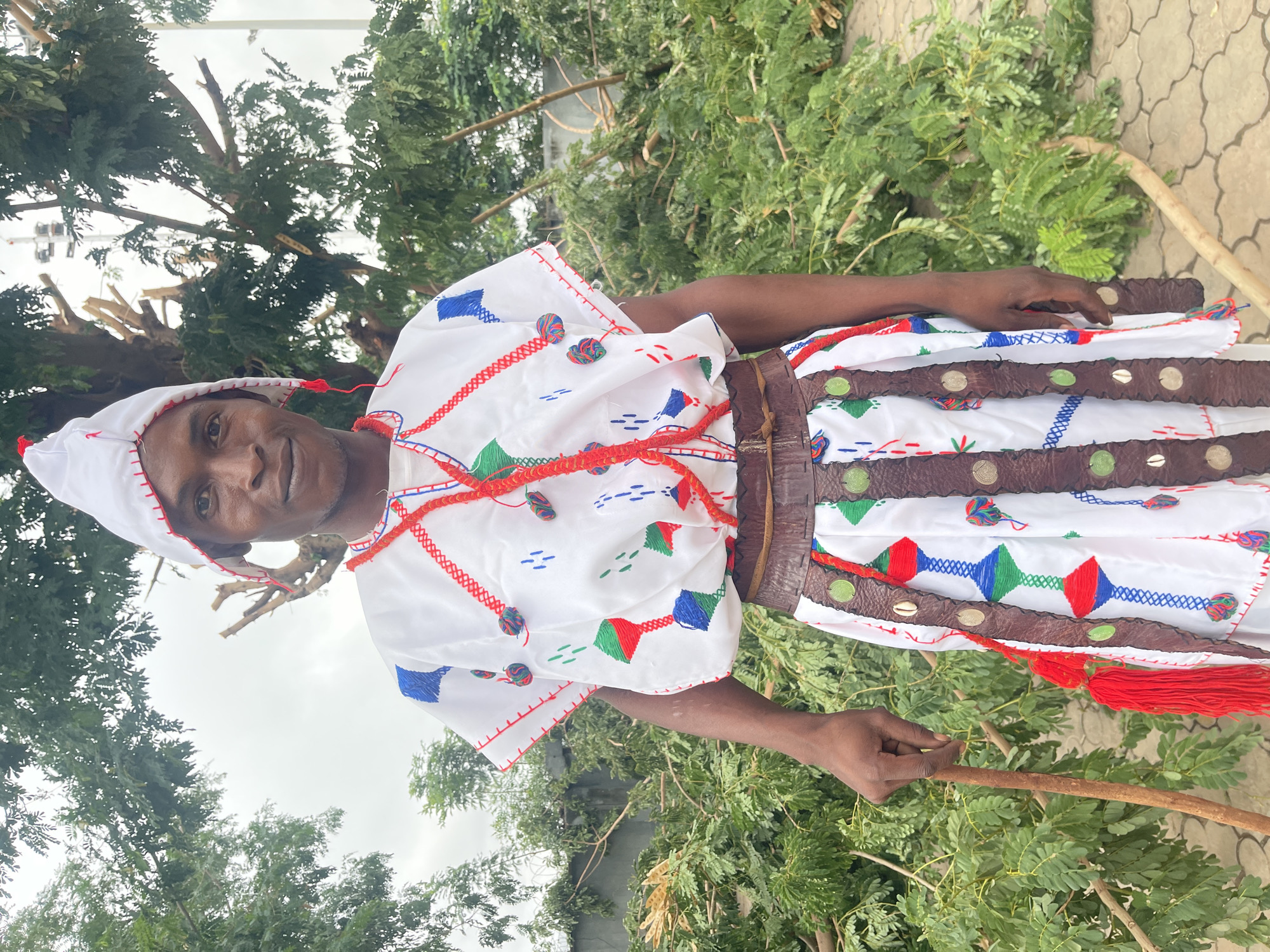 A man stands outdoors, wearing a white tunic with red, blue, and green embroidery, a leather belt, and a white conical hat. He holds a wooden stick in his left hand. The background consists of trees and branches, with a paved ground and a wall visible. The man appears to be in a yard or open space with fallen vegetation, possibly indicating recent storm damage or landscaping. The setting is likely in Takatuku Madorawa, Nigeria.