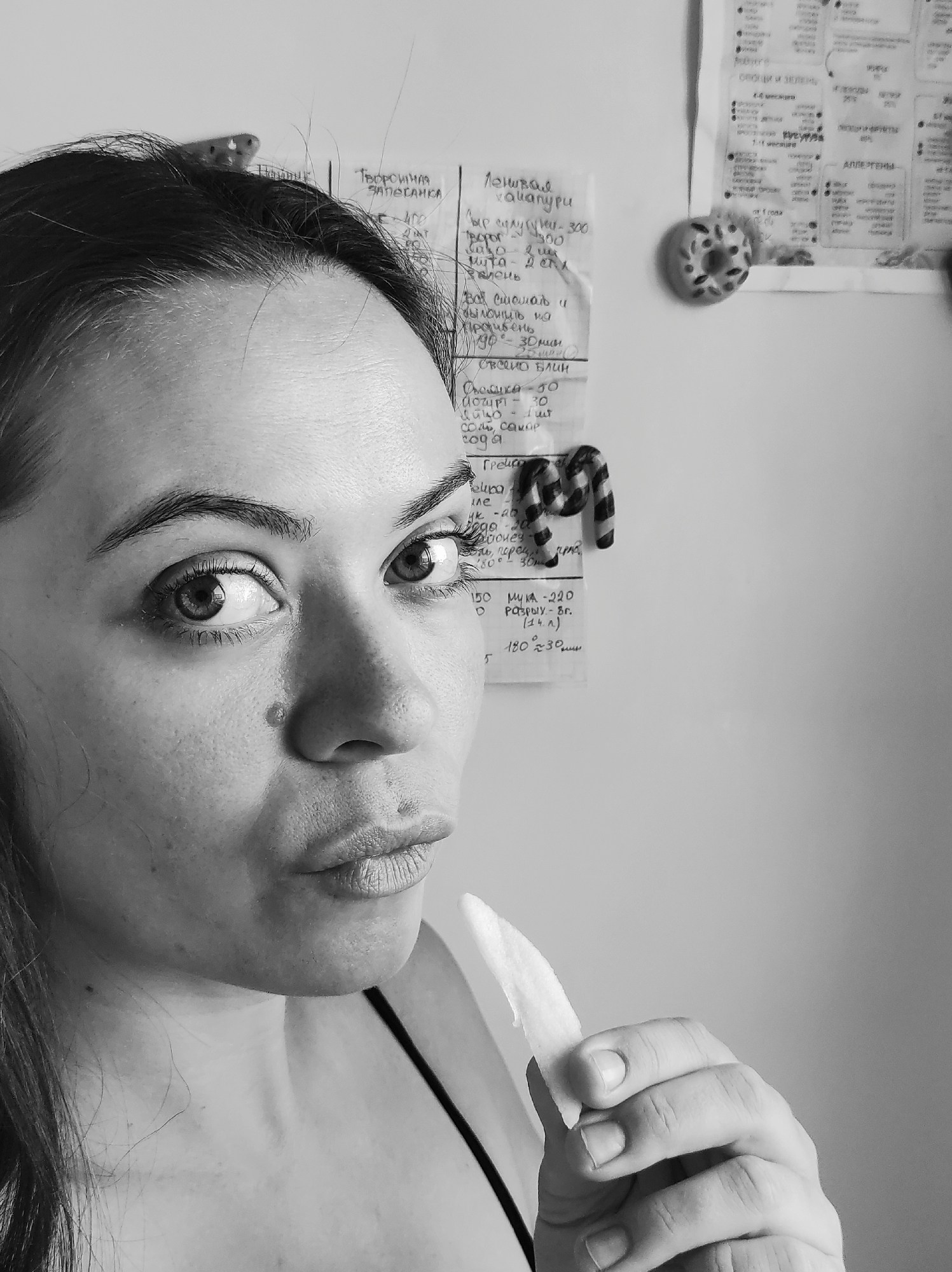 A grayscale photograph depicts a woman indoors. The woman is positioned close to the camera, with her face taking up a significant portion of the frame. She is holding what appears to be a white slice of food, possibly a vegetable, close to her mouth, but not eating it. Her gaze is directed slightly upwards and to the left. The background features a wall covered with notes written in what appears to be Cyrillic script, along with a decorative donut and a striped Christmas candy cane. The location is likely Chaikovsky, Russia, given the Cyrillic writing. The lighting appears to be ambient and diffuse.