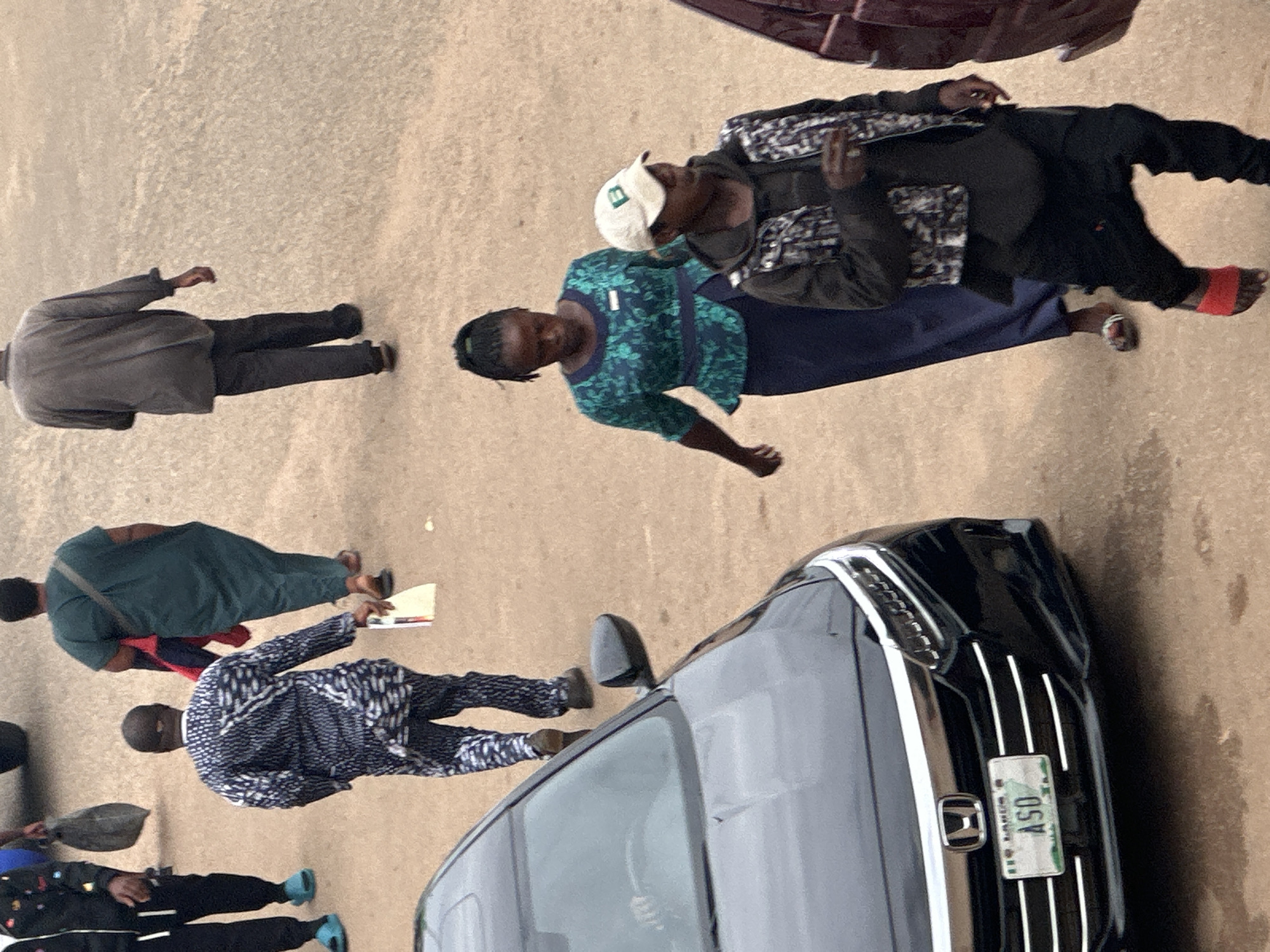 The image shows several people walking on a paved surface, possibly a street, in Bukuru, Nigeria. In the foreground, a dark-colored car is parked, with its license plate visible, reading "ASO". To the right of the car, two individuals are walking. One is wearing a green and blue patterned shirt and a dark blue skirt, while the other, wearing a cap and a patterned vest over a dark hooded jacket, walks ahead. Further back, a man in a dark, patterned traditional outfit walks away from the camera, holding a document. Another person in a dark grey outfit walks in the background. The ground appears to be a mix of dirt and pavement, with scattered debris. The lighting suggests it is daytime.