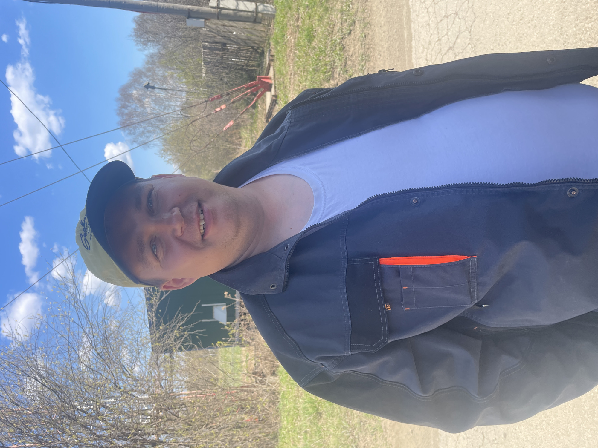 A man stands outdoors in Поедуги, Russia, wearing a white t-shirt and a dark, possibly navy blue, jacket with visible pockets, one with an orange accent. He is also wearing a baseball cap with a possible logo. The background includes a blue sky with scattered clouds, trees, grass, and parts of buildings. The ground appears to be a paved road or path.