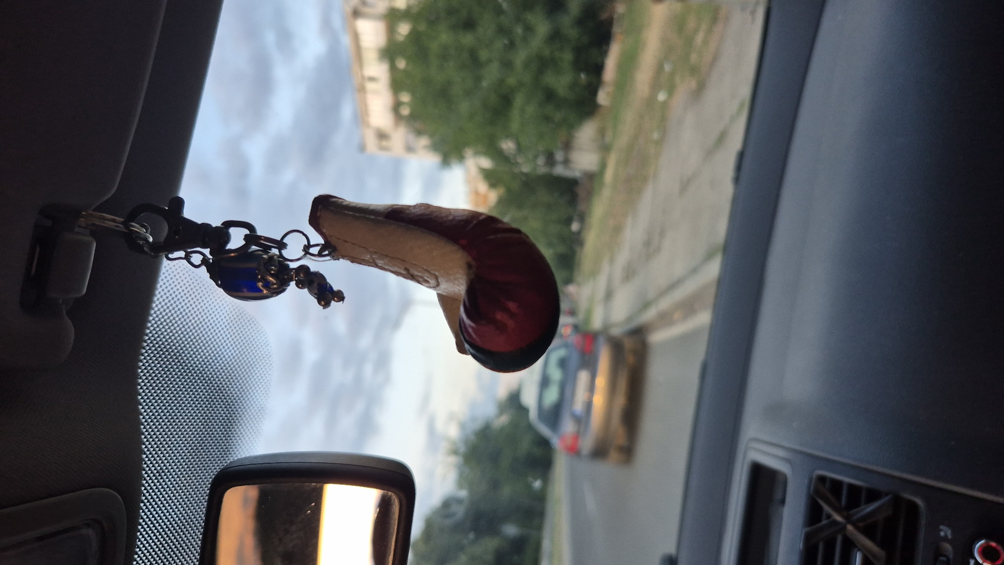 The image is taken from inside a car, looking through the windshield. A keychain with a blue decorative bead and a miniature boxing glove is hanging from the rearview mirror. The boxing glove is red and beige. Outside the car, a grey sedan is driving on a street in Lovech, Bulgaria. The sky is cloudy. The dashboard of the car is visible in the foreground.