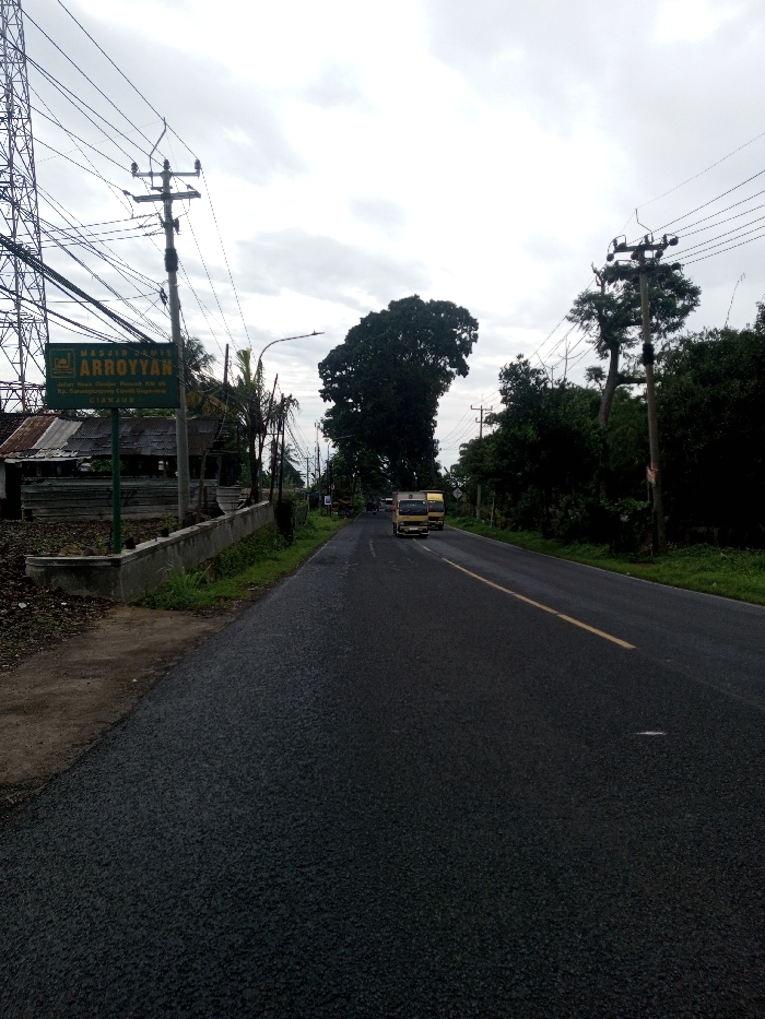 The scene depicts a straight asphalt road under an overcast sky. The road surface appears wet and features a single solid yellow line on its right side, defining a single lane of travel in the visible direction. In the distant center, two light-colored trucks are observed in motion, traveling away from the viewer.
On the left side of the road, a green rectangular sign is prominently displayed, reading "MASJID JAMI ARROYYAN" at the top. Below this, smaller text specifies the address: "Dusun Ciwiru, Parereja, RT.04 RW.01 Ds. Kertamulya, Kec. Lakbok CIAMIS". This indicates the location is Ciamis, Indonesia, which contradicts the requested location of Kamji Na-Yekha, Bhutan. Behind the sign and to its left, a structure with a corrugated metal roof and weathered wooden walls is partially visible. A tall metal lattice tower stands on the far left, connected to multiple utility poles by numerous overhead wires that traverse the scene. A low concrete barrier or retaining wall separates the road from the ground on the left, which consists of dirt and sparse vegetation.
The right side of the road is bordered by a dense line of green foliage and various trees, some of which are tall. Several utility poles with overhead wires are also present along this side. The sky above is uniformly gray with heavy cloud cover. No individuals are visible in the image.
