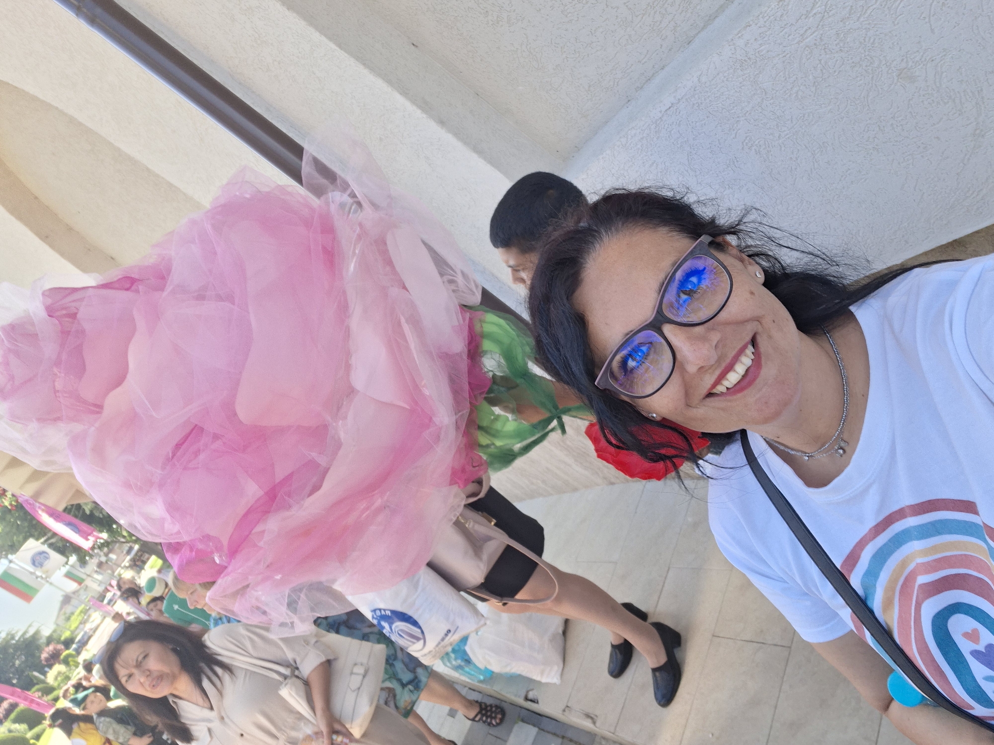 A selfie-style shot depicts a woman with dark hair and glasses smiling, with a crowd and a large pink decoration visible behind her. The woman wears a white t-shirt with a rainbow design and a crossbody bag. The background includes people in a public setting, with Bulgarian flags visible, suggesting a cultural event or celebration. A large, fluffy pink decoration is prominent on the left side of the image. The location is identified as Pavel Banya, Bulgaria.