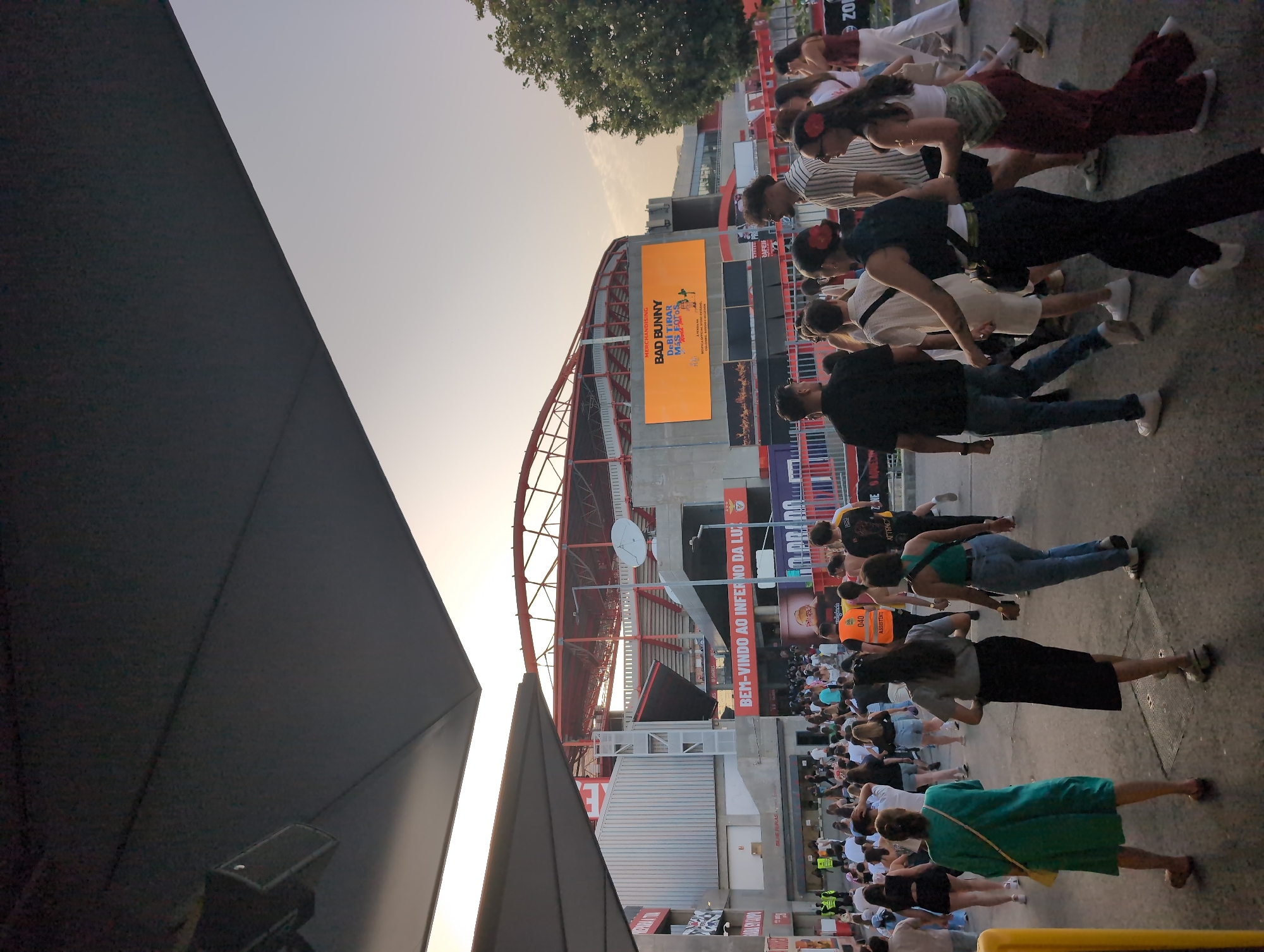 The image shows a crowd of people standing outside Estádio da Luz in Lisbon, Portugal. Many people stand around, dressed in a variety of clothes. There is a large screen showing an advertisement for Bad Bunny, and the stadium's red metal framework is visible. The scene appears to be an outdoor event or gathering before a concert or game. A dark, angular structure is on the left side of the image.