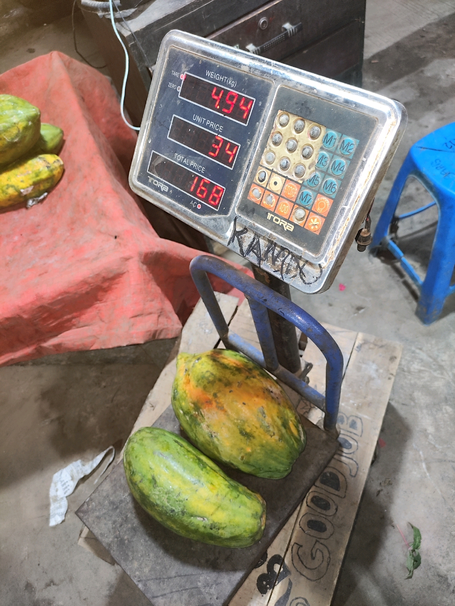 The image depicts a scene in a market environment, potentially in Karawang, Indonesia, based on the graffiti. At the center is a digital weighing scale with a blue metal frame holding a flat weighing platform. On the platform, two papayas are placed, one predominantly green and the other showing signs of ripeness with yellow and orange patches. The scale displays a weight of 4.94 kg, a unit price of 34, and a total price of 168. The scale's keypad area is visible, along with some worn buttons and a "TORA" logo. To the left of the scale, a red cloth is spread out, with a few more papayas or similar fruits piled on it. In the background, a dark wooden cabinet or counter is visible. To the right, a bright blue plastic stool is present. The ground appears to be concrete. Graffiti is visible on the scale, including the name "KANIX" and the phrase "GOOD JOB" scrawled on a wooden plank beneath the scale. The overall impression is that of a market stall where produce is being weighed and priced.
