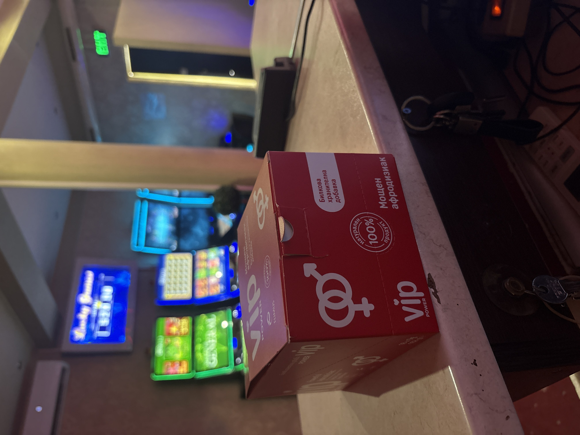 The image depicts the interior of a dimly lit establishment, possibly a casino or a gaming hall, in Karlovo, Bulgaria. In the foreground, a red cardboard box with the "VIP POWER" logo is placed on a counter. The box features symbols representing male and female genders, and text in Bulgarian that translates to "Herbal food supplement" and "Potent aphrodisiac." To the left and in the background, several slot machines are visible, their screens displaying colorful graphics and text. An air conditioning unit is mounted on the wall above the slot machines. A green "EXIT" sign is illuminated on the right side of the image, indicating a fire exit. The overall ambiance suggests a commercial establishment operating in the late hours.