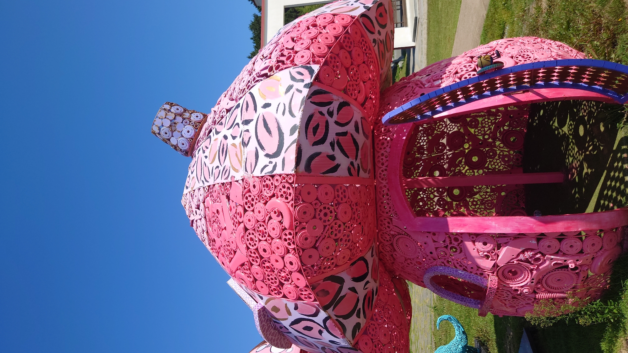 The image shows a large, pink, mushroom-shaped sculpture made of recycled metal parts, possibly gears and other mechanical components. The sculpture is located outdoors in a grassy area with a clear blue sky overhead. In the background, there is a building with white walls and a white roof, and some trees. The sculpture has an open entrance and appears to be a whimsical art installation. This sculpture is located at the Ulsan Grand Park, in the city of Ulsan, South Korea.