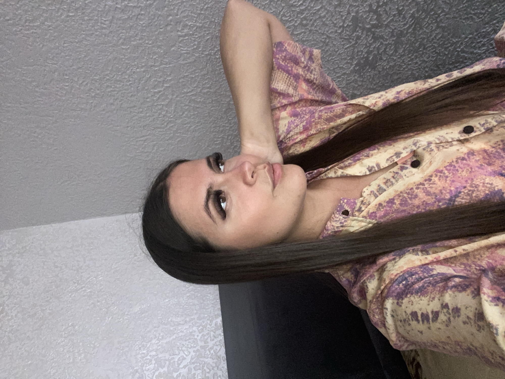 A woman with long, dark hair and dramatic false eyelashes poses with her right arm behind her head. She is wearing a button-up shirt with a psychedelic pattern of purple, pink, and yellow. The background is a textured gray wall. The image appears to be taken indoors. There is no indication of the city of Chalda, Russia, in the image.