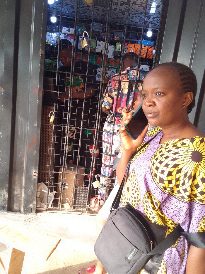 The image displays an outdoor commercial scene in Nkwelle Ezunaka, Nigeria. In the foreground, an adult female with braided hair, wearing a purple garment with yellow and white geometric patterns, holds a dark-colored smartphone to her right ear, engaged in a phone call. She carries a black cross-body bag slung over her left shoulder. Behind her, a dark-colored metal security gate, constructed of a grid pattern, stands partially open, secured by two visible gold-colored padlocks.
Behind the gate, a retail shop interior is visible. Two adult males are present inside. The male on the left wears a black t-shirt with a circular logo, while the male on the right wears a multi-colored patterned shirt, largely facing away from the camera. The shop's shelves are stocked with various items, primarily electronic accessories in distinct colored packaging. A bright white LED bulb illuminates the shop's interior from above. The ground in the immediate foreground is a light-colored paved surface, with a wooden plank visible on the left. Bright daylight illuminates the exterior portion of the scene.