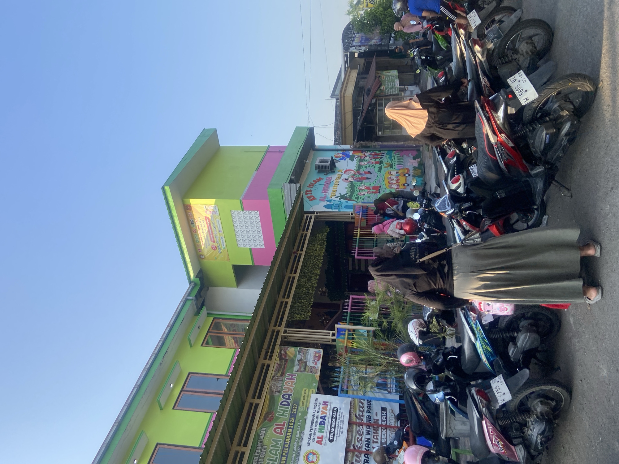 This is an eye-level, medium shot of the front of a colorful building, likely a school, in Ngampelsari, Indonesia. The building is painted in vibrant shades of lime green, pink, and yellow, with a green-tiled roof. Several banners are displayed on the front of the building. In the foreground, a row of parked motorcycles occupies the paved area. A woman in a black long-sleeved shirt and a long, khaki skirt with a beige headscarf stands with her back to the camera, facing the motorcycles. She is wearing light-colored sandals. Behind her, other individuals, some also wearing headscarves, are visible in smaller numbers. The sky is clear and blue.