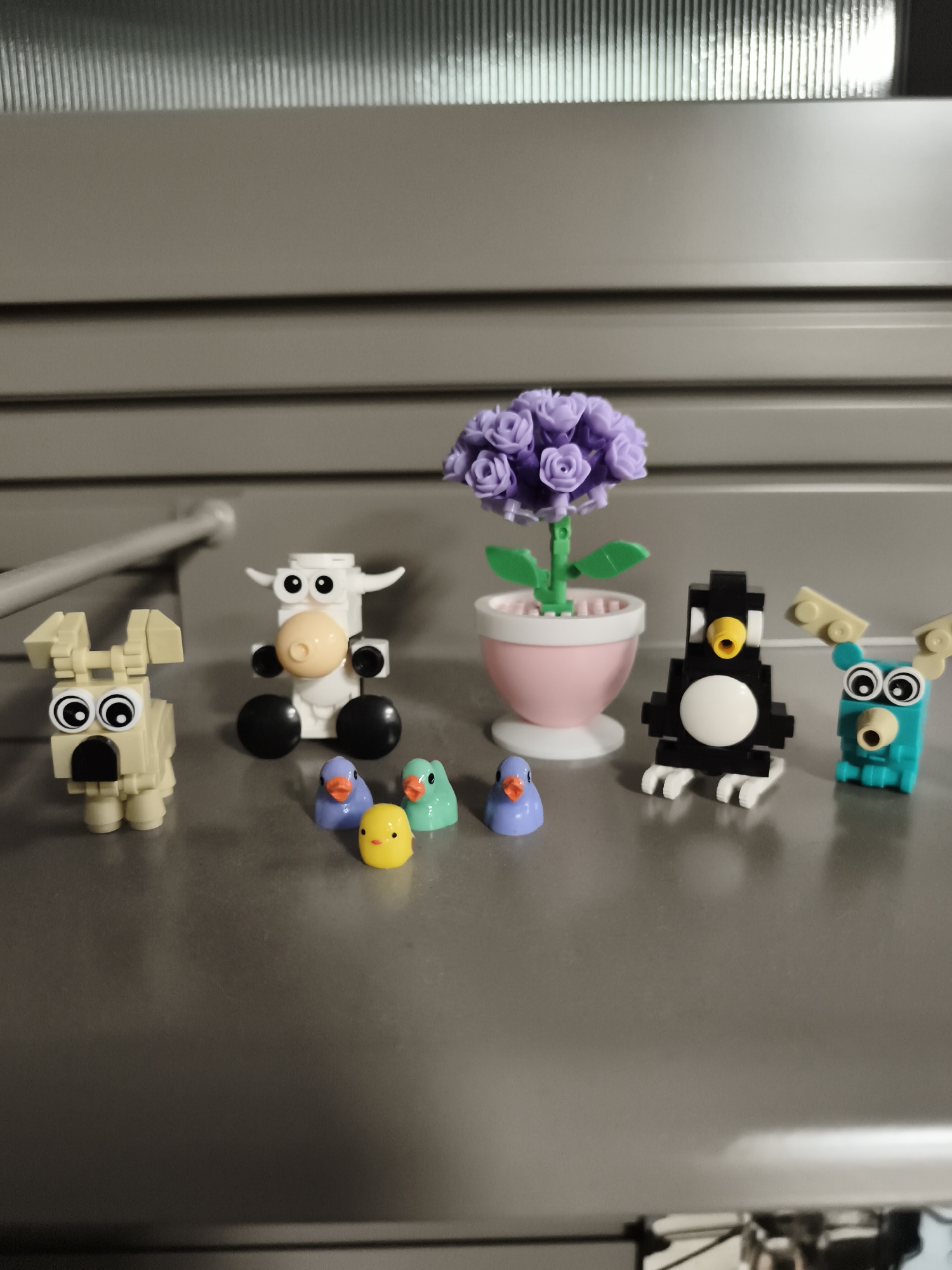 The image shows a collection of small, brightly colored toy figures arranged on a metallic surface. On the left is a tan-colored dog-like figure with large, round eyes and an open mouth. To its right is a white cow figure with black eyes, a light orange nose, and black wheels for feet. In front of the cow are three small bird-like figures: one yellow, and two in shades of blue and green. Behind them is a pink pot with a purple flower arrangement consisting of multiple small roses. To the right of the flower pot is a black and white penguin figure with a yellow beak and white feet. Further to the right is a teal-colored creature with large eyes and tan antlers. The background consists of horizontal, ribbed panels in a light brown color. There is no indication of a location in the city of Columbia, United States of America.