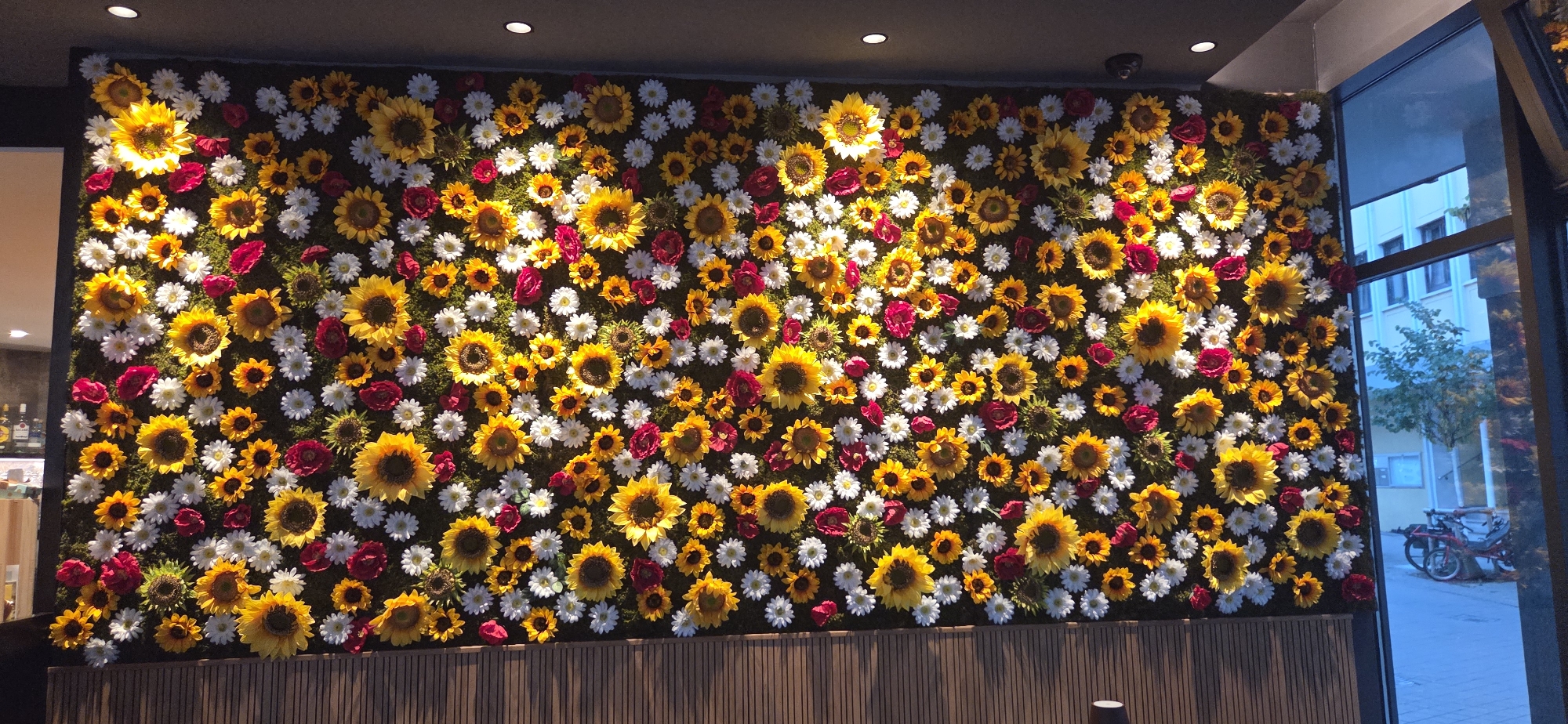 The image shows a vibrant wall covered in a dense arrangement of artificial flowers, predominantly sunflowers in various sizes, interspersed with white daisies and red roses. The floral display forms a textured, naturalistic backdrop against a dark wall. The bottom of the wall is bordered by a ribbed wooden panel. To the right, a large window reveals an outdoor scene with a light-colored building and a tree, suggesting an urban setting. Parked bicycles are visible outside the window. The lighting within the indoor space appears to be artificial, with some spotlighting on the floral wall. This scene is located at the entrance of "Das Bootshaus," a restaurant in Friedrichshafen, Germany, as indicated by the distinctive sunflower wall art.
