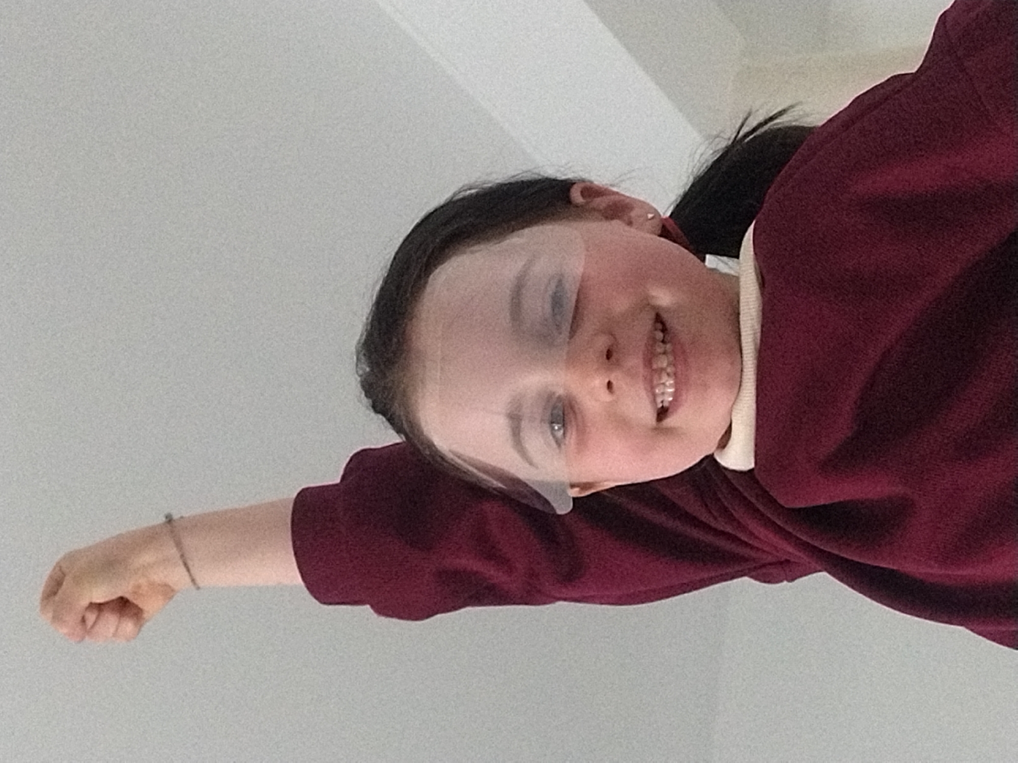 A fair-skinned young female with dark hair pulled back into a ponytail is depicted in a photograph. She is wearing a maroon sweater over a collared shirt. Her right arm is extended forward, with a closed fist. There is a bracelet visible on her right wrist. A translucent object partially obscures her eyebrows. She has braces on her teeth and is smiling. The background is a plain white or light grey color. The photo's orientation is rotated, with the subject appearing to be lying or leaning at an angle. There is no evidence to suggest the image was taken in Crauthem, Luxembourg.