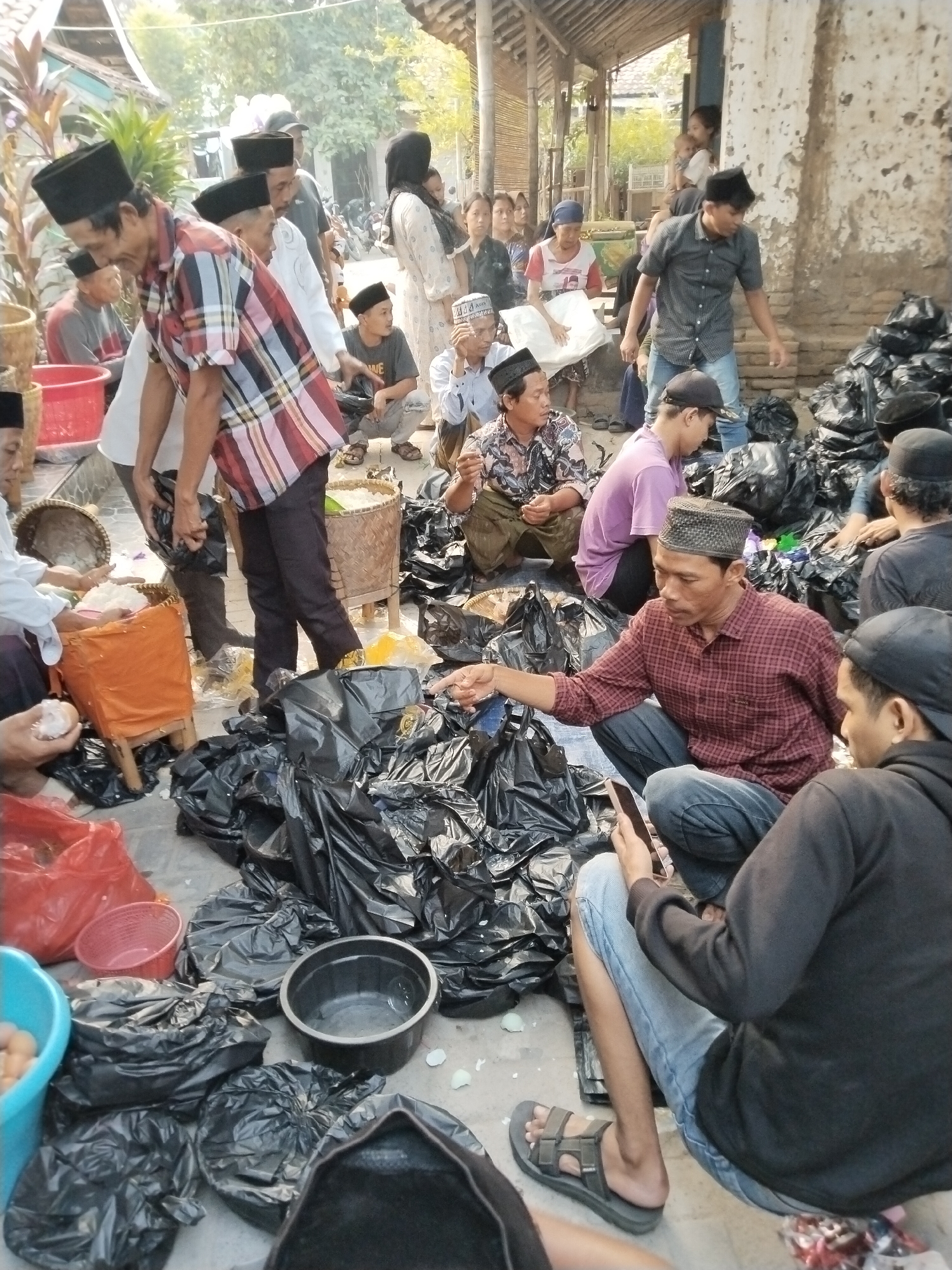 The image depicts a street scene in Serang, Indonesia, likely near a mosque or a traditional market, judging by the attire of the people and the presence of food distribution. A group of men and women, most of whom are wearing traditional Indonesian Islamic attire such as peci hats and modest clothing, are gathered. Several individuals are engaged in distributing or receiving food items, which appear to be rice and possibly eggs, packaged in black plastic bags and baskets. Some people are sitting on the ground amidst the distributed items, while others are standing. The environment shows a slightly unkempt, outdoor setting with a building in the background, suggesting an urban or semi-urban area. The overall atmosphere appears to be one of communal gathering and food distribution, possibly for charity or a religious event.