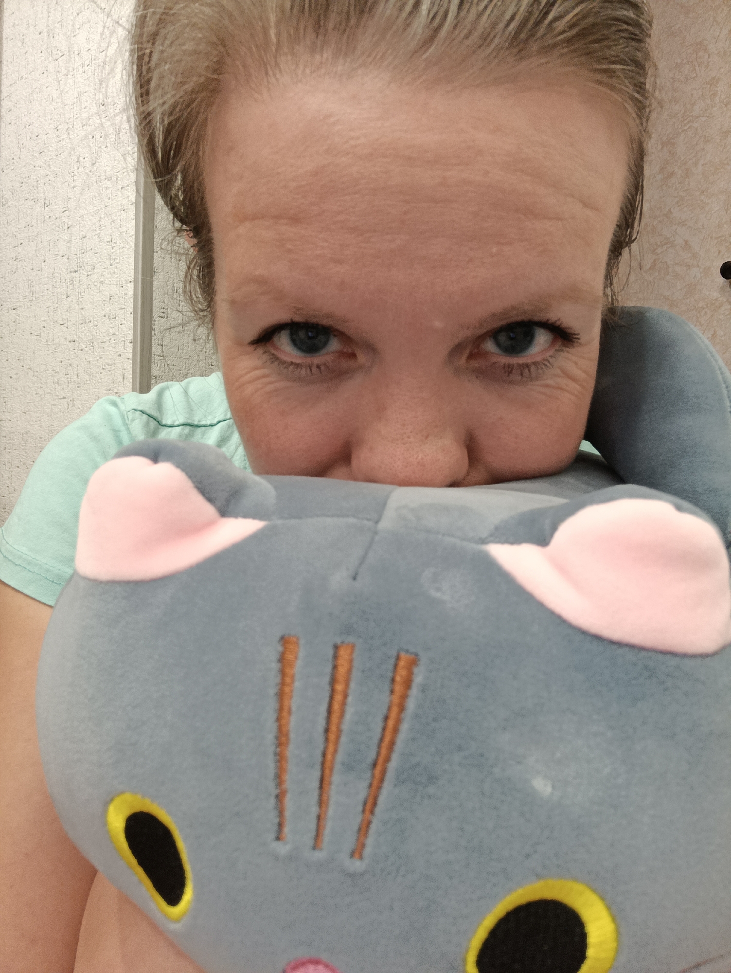 A close-up, eye-level shot features a woman peeking over a plush toy, looking directly at the camera with an intense gaze. The woman has light skin and light-colored hair, pulled back, revealing her forehead with visible wrinkles and her blue eyes. She wears black eyeliner on her upper lash line, and crow's feet are visible around her eyes. Her nose is partially visible just above the toy. She is wearing a light mint green t-shirt.
The prominent object in the foreground is a grey or blue plush toy, resembling a cartoon cat or animal. It has distinct pink inner ears, large yellow and black oval-shaped eyes, and three vertical orange stitched lines on its forehead. The toy covers the lower part of the woman's face, from her mouth downwards.
The background suggests an indoor setting. To the left, there is a light-colored, textured wall, possibly white or off-white, with a vertical line that could be a doorframe or paneling. To the right, another section of wall appears to be a warmer, light beige or cream color with a different texture.
The overall impression is a playful or curious moment, possibly a selfie. There is no visible text in the image. The location context provided is Chaikovsky, Russia.