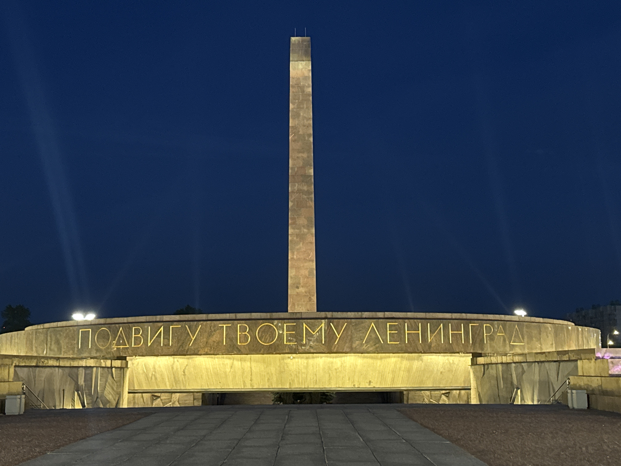 This image captures the Monument to the Heroic Defenders of Leningrad at Victory Square (Ploshchad Pobedy) in Saint Petersburg, Russia, during twilight. A tall, slender, tapering obelisk, constructed of light-colored stone, stands centrally, illuminated with warm artificial light against a deep, uniform blue sky. At its base is a large, circular, multi-tiered structure made of concrete or stone. The inscription "ПОДВИГУ ТВОЕМУ ЛЕНИНГРАД" (To Your Heroic Deed, Leningrad) is prominently displayed in glowing golden Cyrillic capital letters on the upper curved surface of this base. Below the inscription, a recessed area serves as an entrance, featuring textured, angular walls that are brightly lit from within. The monument is devoid of people. In the foreground, a paved walkway of grey rectangular tiles extends towards the monument, bordered by darker ground material. Scattered artificial lights are visible on and around the circular structure, and faint light rays emanate from the left side of the frame. Dark tree silhouettes are visible on the left, and faint distant building lights are present on the far right.
