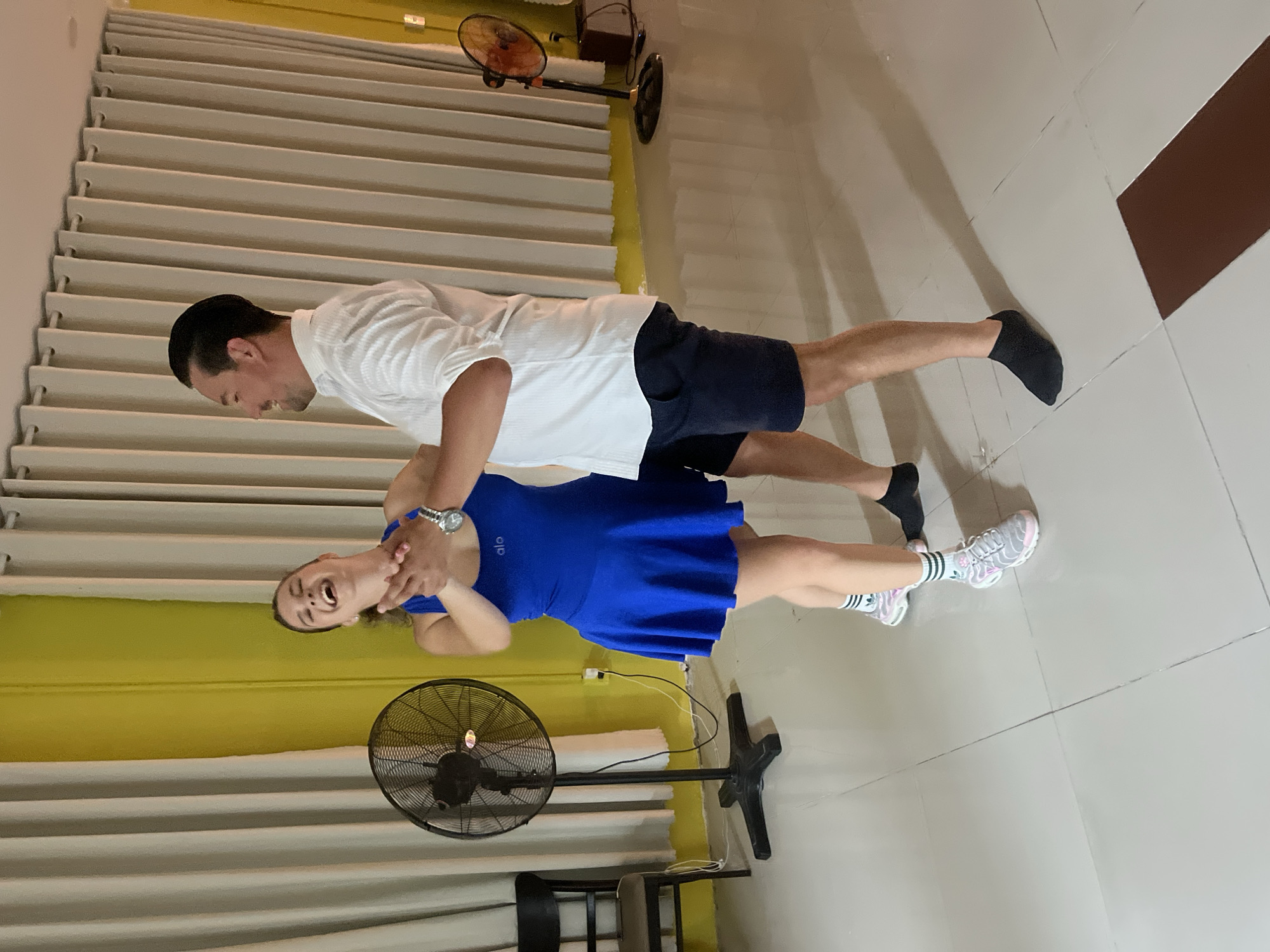 This media file shows a man and a woman in what appears to be an indoor dance studio or a similar recreational space. The man, wearing a white polo shirt and dark shorts, is positioned behind the woman. He is leaning forward, holding her torso. The woman, in a bright blue athletic dress and athletic shoes, is facing away from the camera and appears to be interacting with the man. Her mouth is open as if she is speaking or laughing, and her hands are near her face.
The setting features a wall with horizontal white blinds or louvers behind the subjects. To the left of the frame, a large black oscillating fan is visible on the floor. In the background, a smaller, older-style orange fan is mounted on a stand. The floor is tiled with light-colored squares. The lighting suggests an indoor environment, and the presence of fans indicates a potentially warm climate, consistent with the location context of Đà Nẵng, Vietnam. There are some visible shadows on the floor, indicating a light source from above and to the side. The overall mood seems to be playful and energetic, possibly during a dance lesson or practice.
The visible text on the woman's dress reads "alo." The scene is shot in a slightly tilted perspective. The time of day cannot be definitively determined from the image, but the indoor setting and artificial lighting make it less dependent on natural light cues.