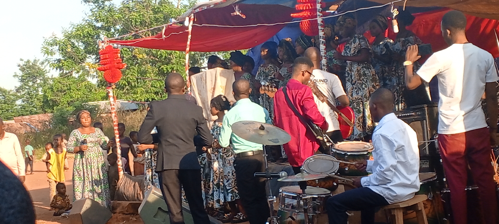 The image shows a musical performance taking place outdoors, possibly in a neighborhood of Kalemie, Democratic Republic of the Congo, judging by the visible landscape and the ethnicity of the people. A group of individuals is positioned on a makeshift stage covered by red and blue tarps. Several women in patterned dresses stand behind a music ensemble. The music group includes a drummer playing a drum kit, several guitarists, and other musicians. A woman in a green and white patterned dress stands with a microphone in front of the stage, likely singing or speaking. People in the audience are positioned around the stage; children stand closer to the speaker. A man in a white shirt with the word "SOUND" printed on the back is partially visible on the stage. The scene appears to be an event of some kind, potentially religious or cultural, given the presence of musicians and the size of the crowd. A decorative red object is visible to the left of the stage. A tree is present in the background.